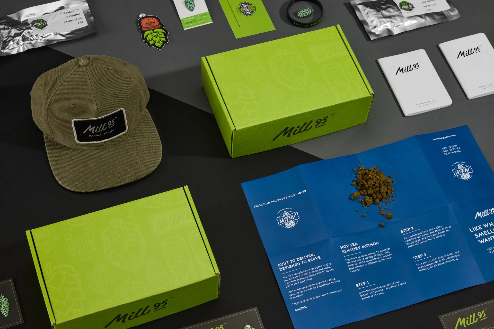 Mill 95 branded merchandise including a hat, green boxes, brochures, and various promotional items displayed on a grey surface.