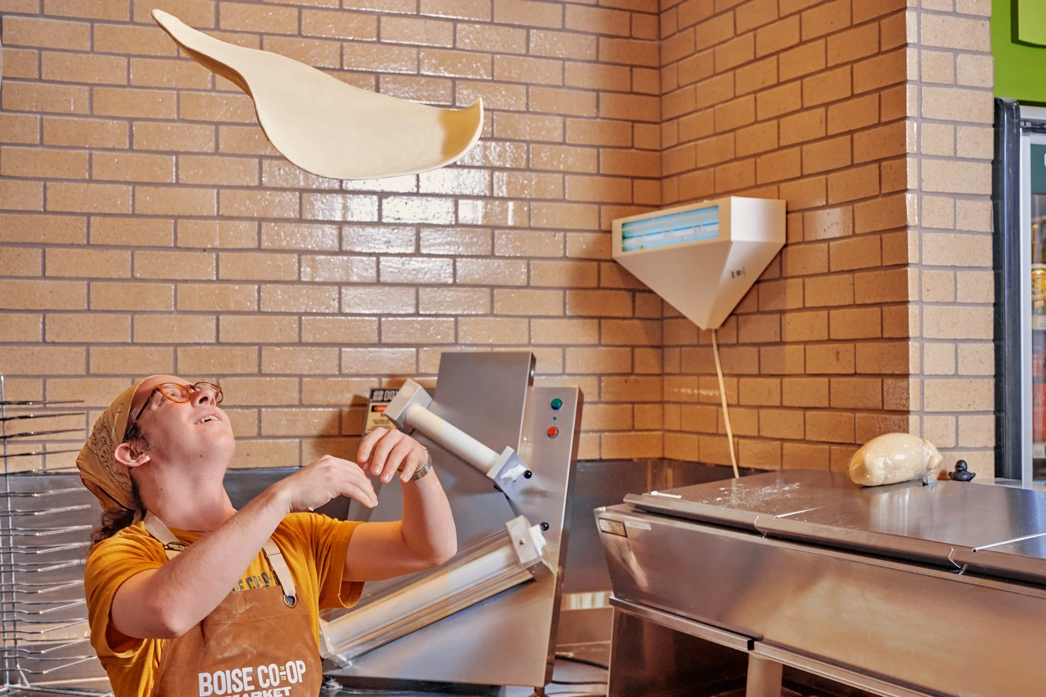 Boise Co-op worker tossing pizza dough in the air inside a bakery prep area.
