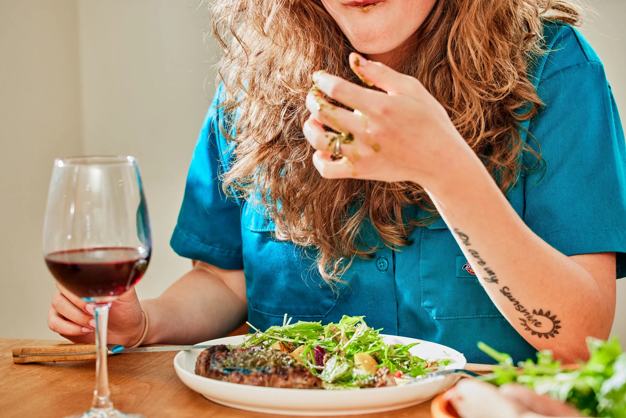 Person in blue shirt eating a salad with a steak, holding food with their hand, and a glass of red wine on the table.