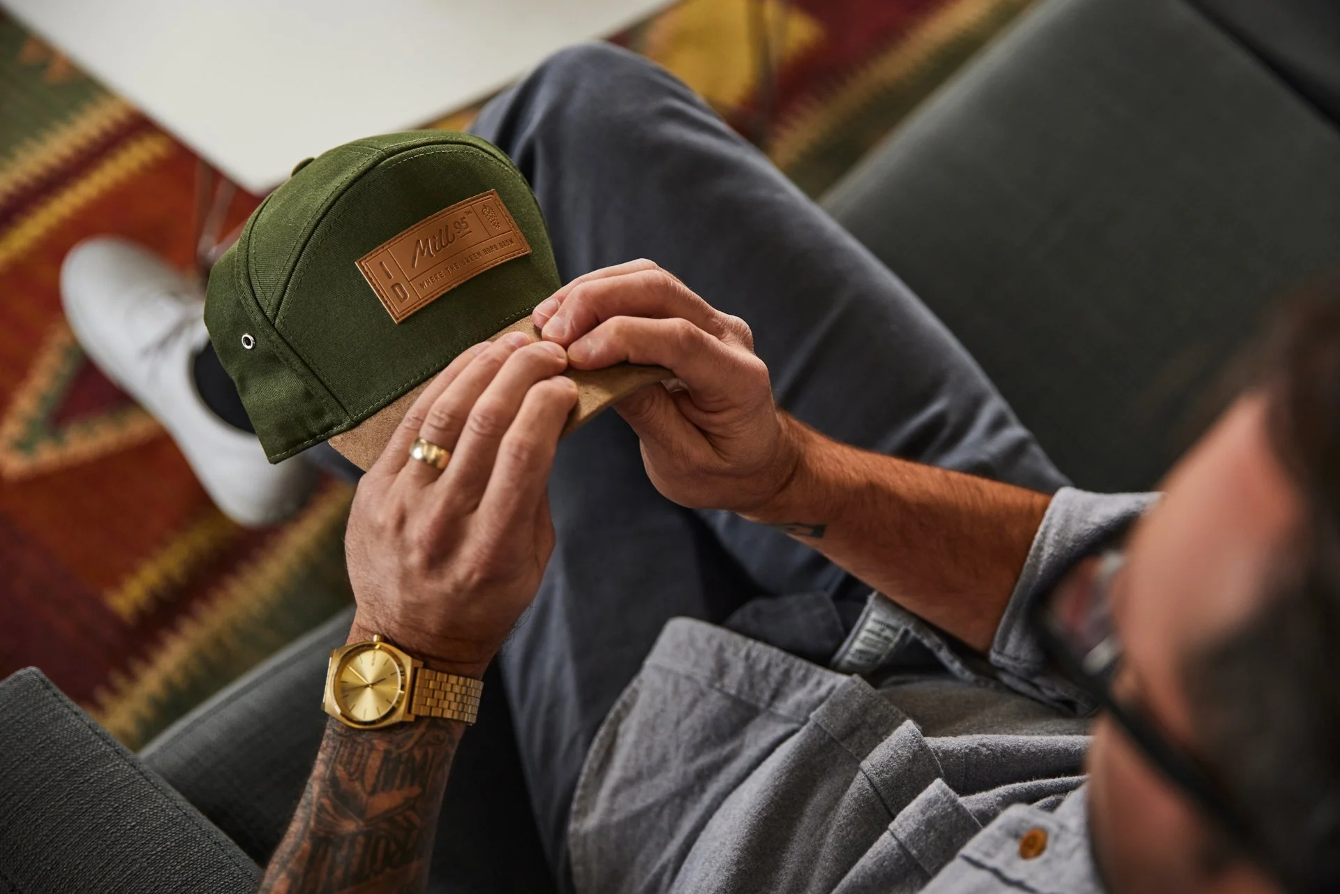 Man holding green cap with brown leather patch, wearing a gold watch and gray shirt.