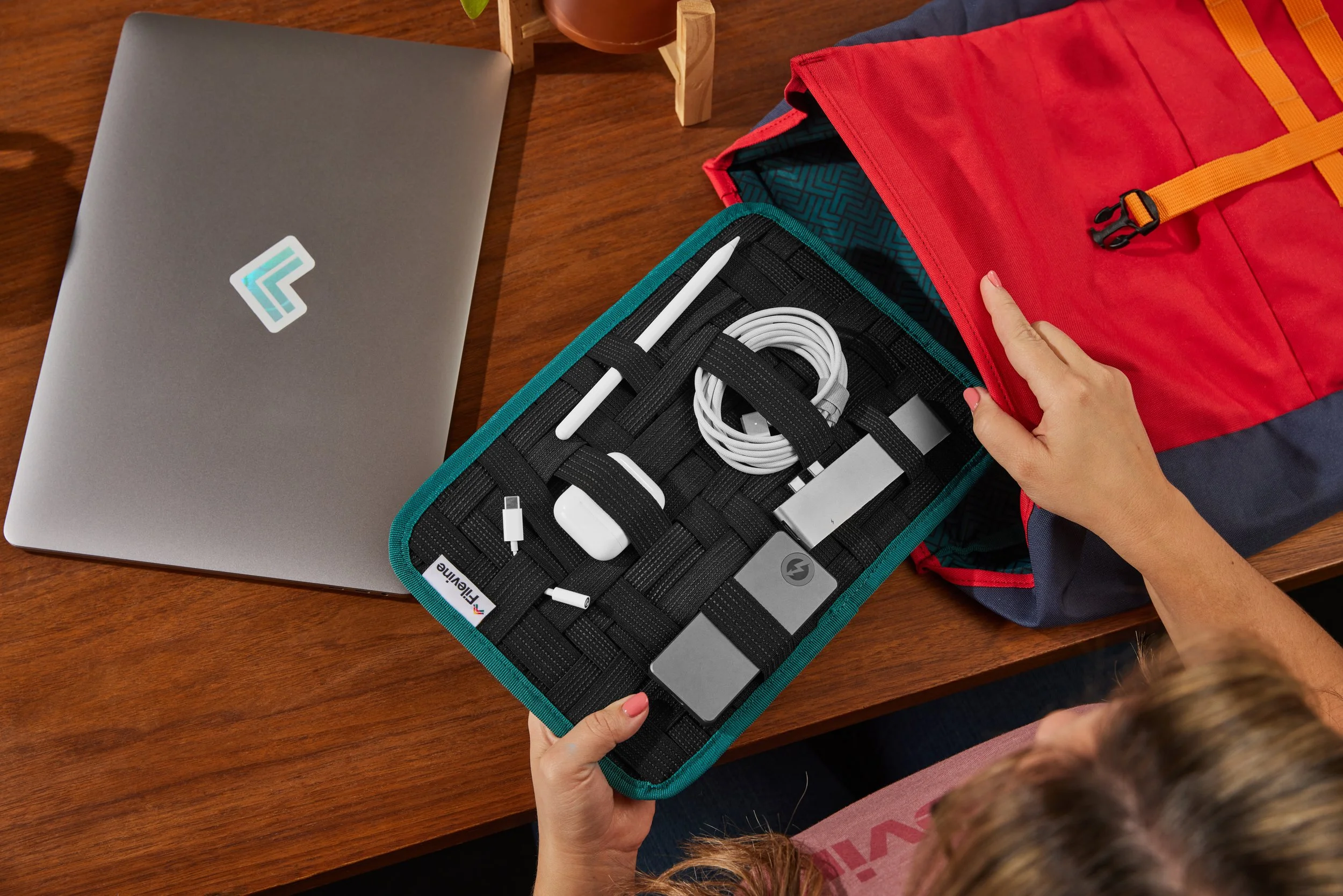 Person organizing a tech organizer with gadgets and cables on a wooden desk next to a laptop and red backpack.