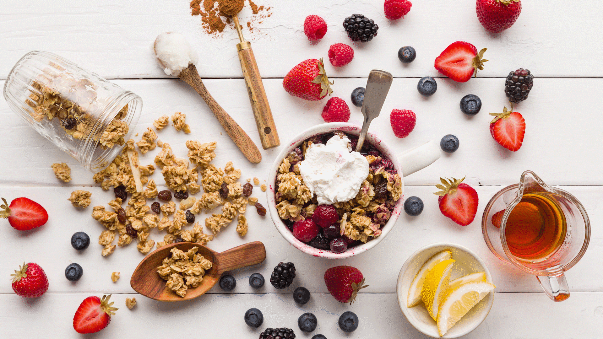 Healthy breakfast setup with granola, fresh berries, sliced strawberries, blueberries, blackberries, raspberries, honey, lemon slices, cinnamon, and yogurt on a white wooden table.