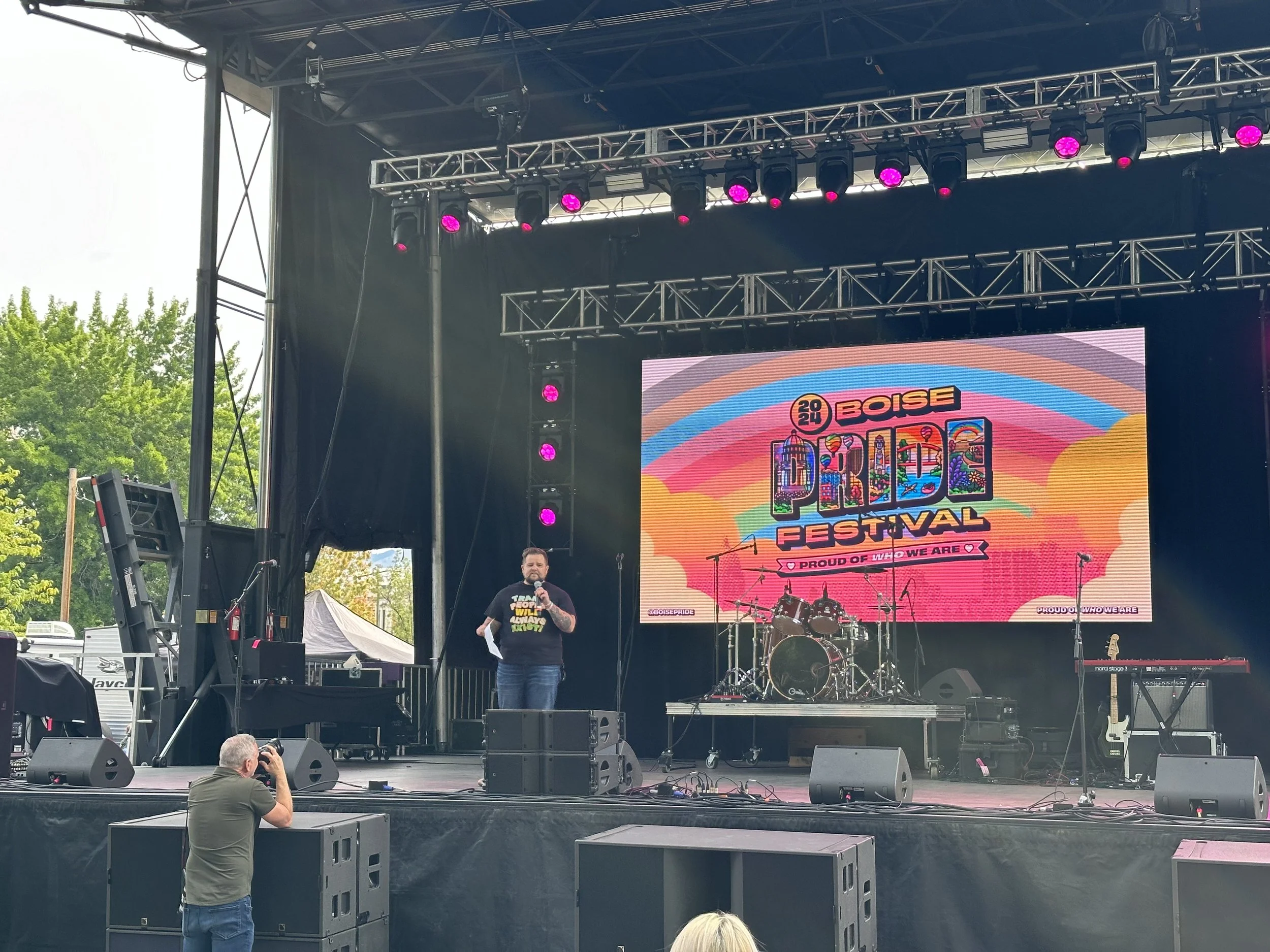 Person speaking on stage at Boise Pride Festival with colorful backdrop, musical instruments, and stage lighting. Photographer in foreground.