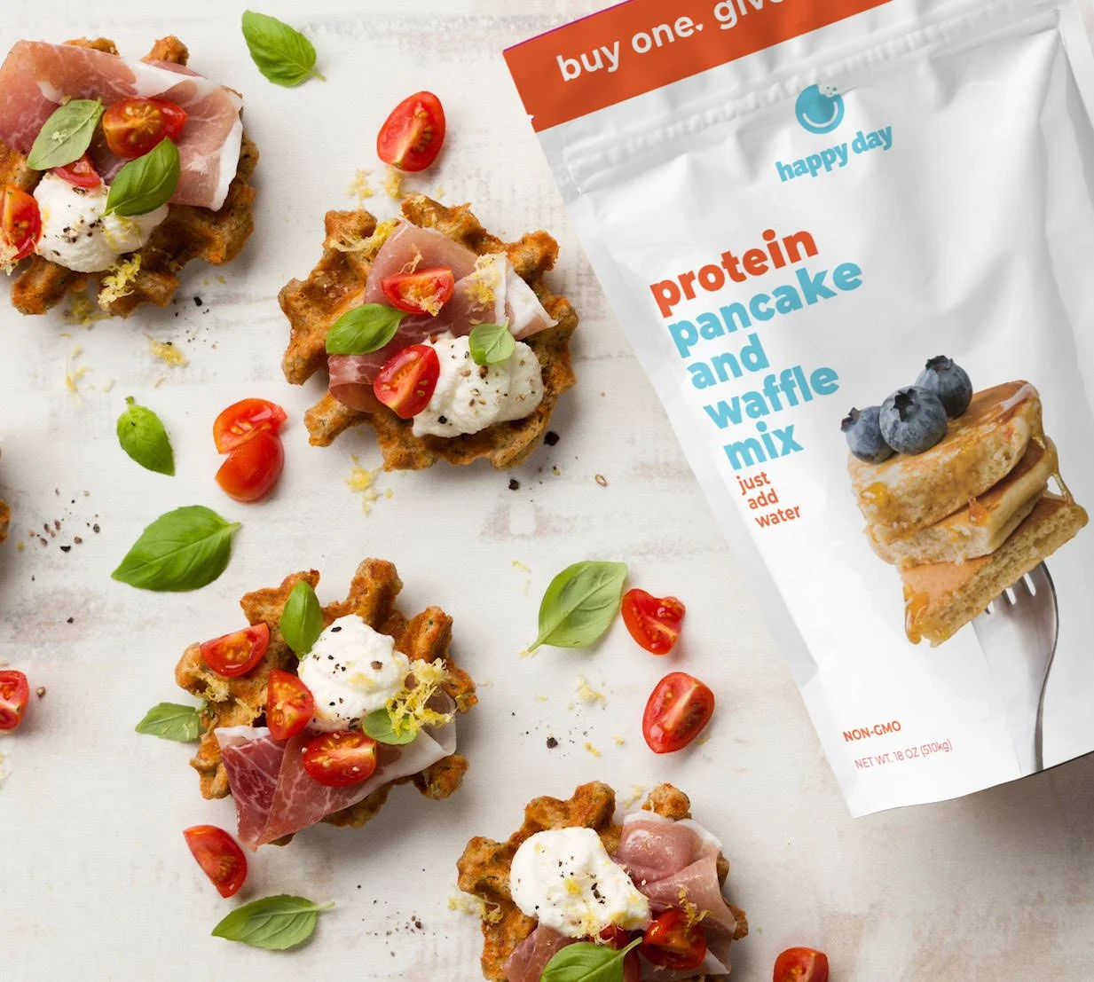 Waffles topped with cream, cherry tomatoes, ham, and basil leaves next to a package of protein pancake and waffle mix.