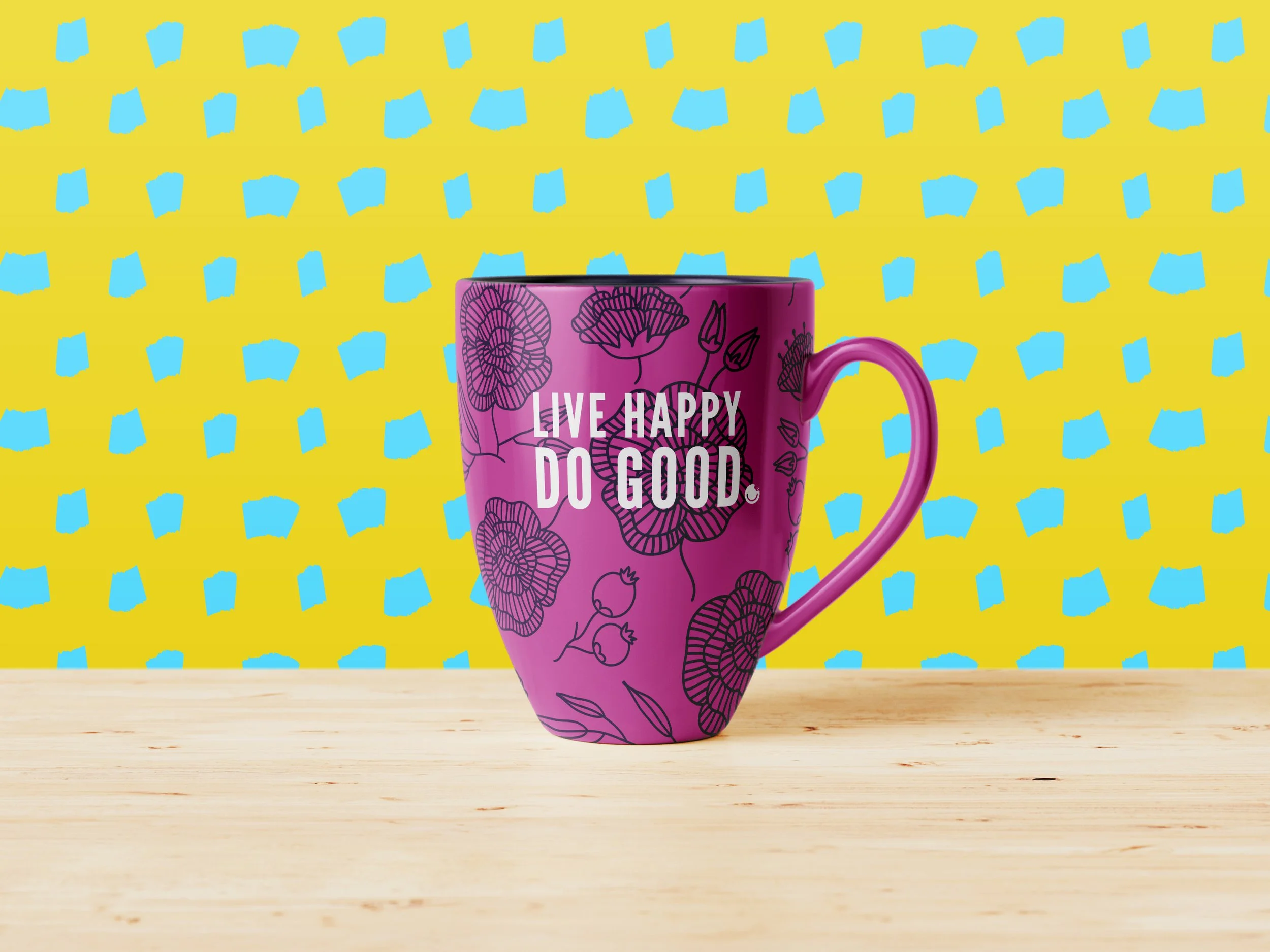 Pink mug with "Live Happy Do Good" text, floral design, on wooden surface; bright yellow and blue patterned background.