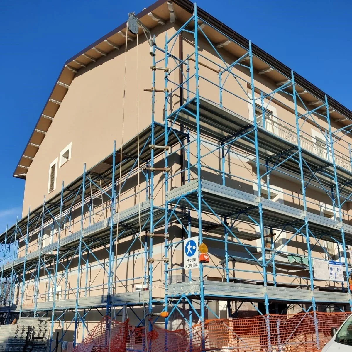 Edificio in costruzione con impalcature blu e segnali di sicurezza, sotto un cielo blu.