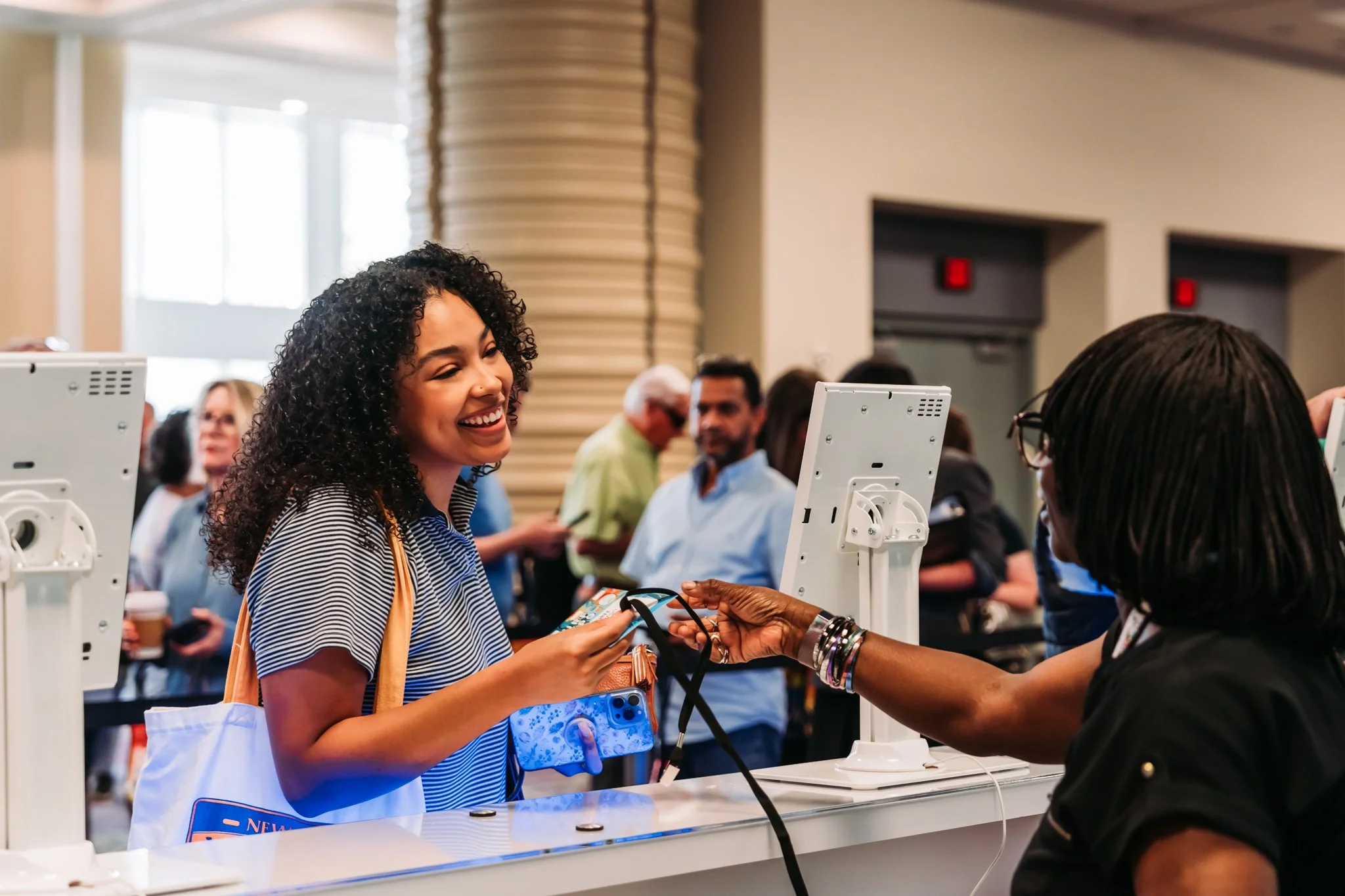 A woman at a conference or event receives a lanyard from a woman behind a registration desk, smiling while others in the background talk and walk around.