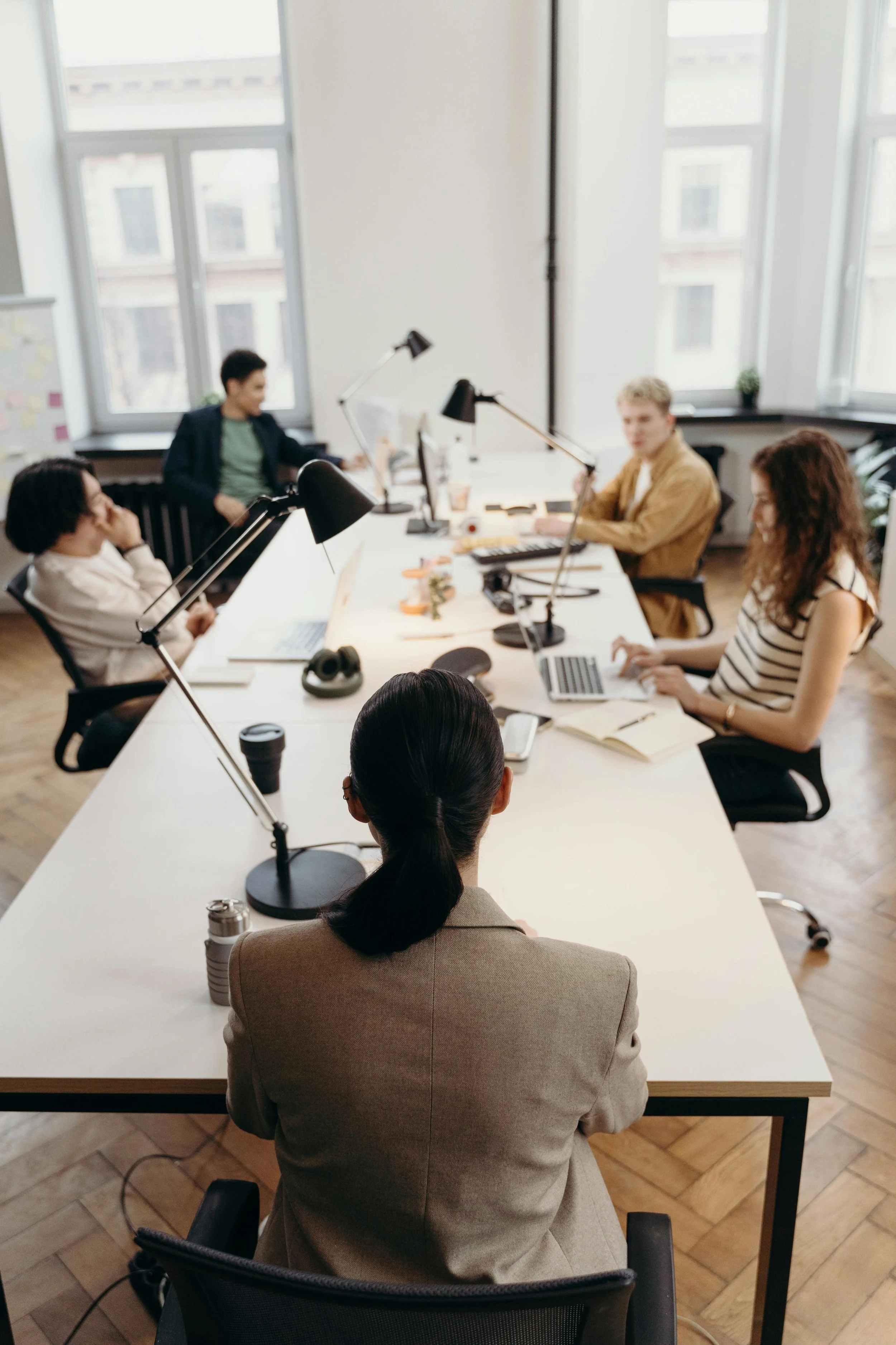 A woman with dark hair in a ponytail sitting at a conference table, attending a meeting with four other people in a bright office with large windows.