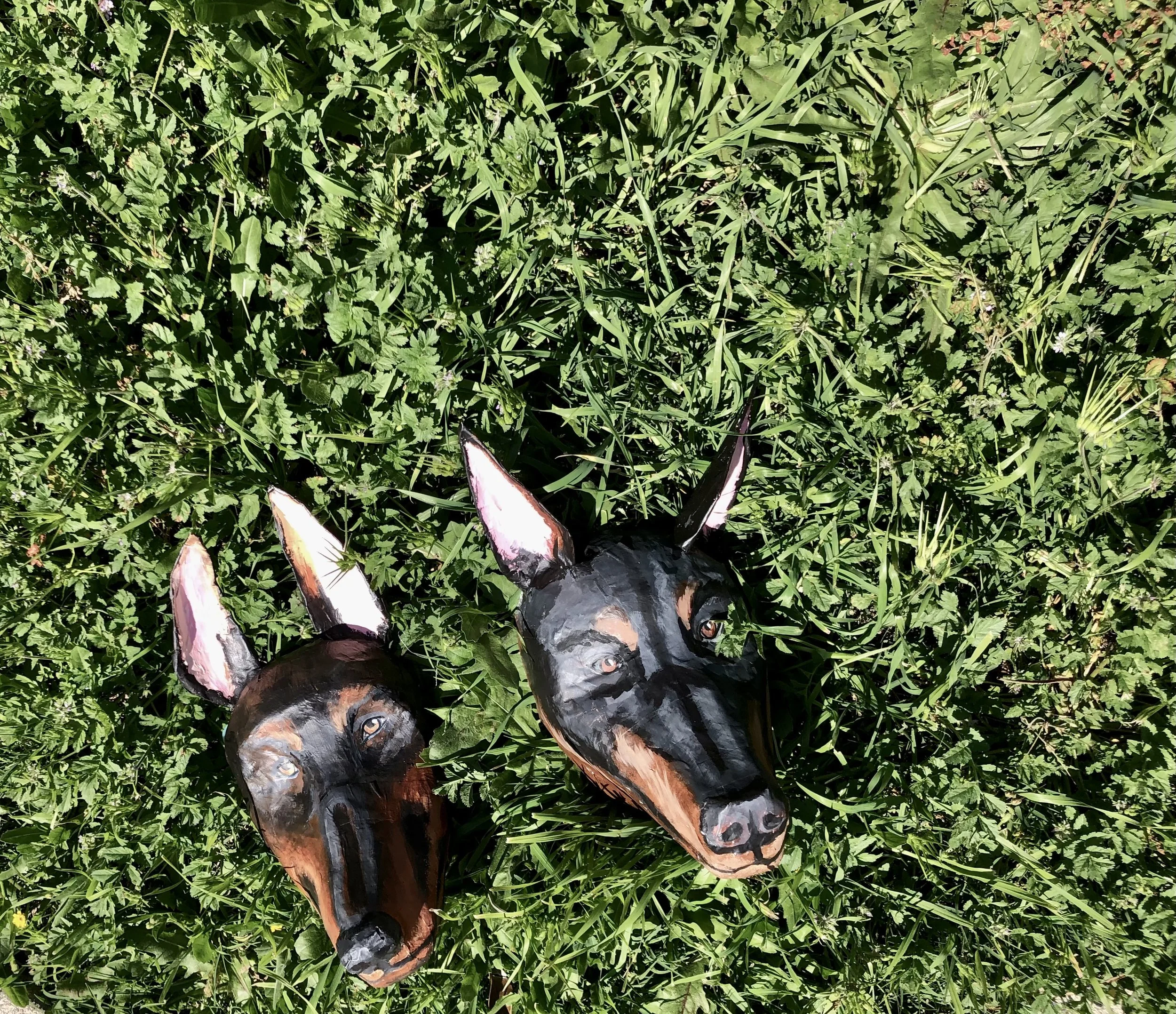 Two handmade dog face masks crafted from paper mache, photographed on grass. Contemporary wearable art masks with playful animal-inspired design, original sculptural pieces by Giana Amantea.