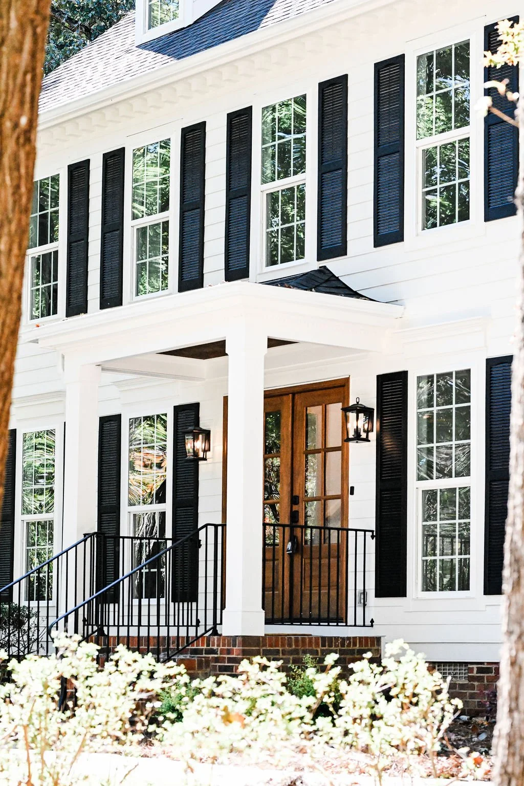 Front view of a white two-story house with black shutters, wooden front door, brick steps, black railing, and outdoor lanterns, surrounded by trees and bushes.