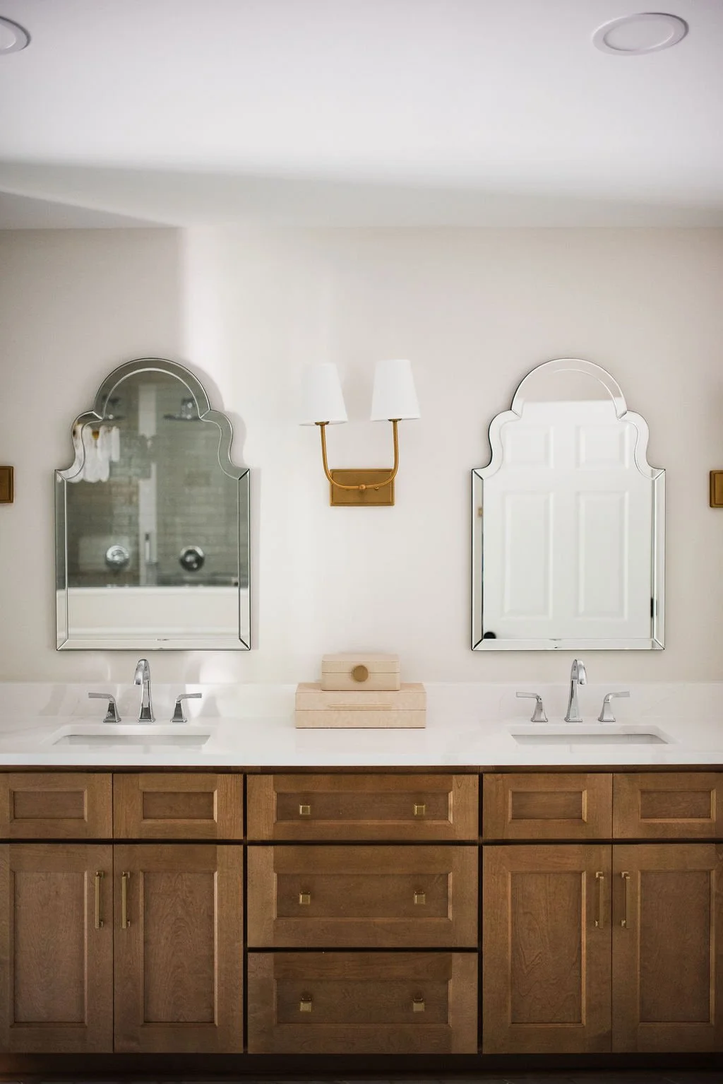 Bathroom vanity with two mirrors, a double sink, and a wooden cabinet. Two wall-mounted light fixtures are placed between the mirrors.
