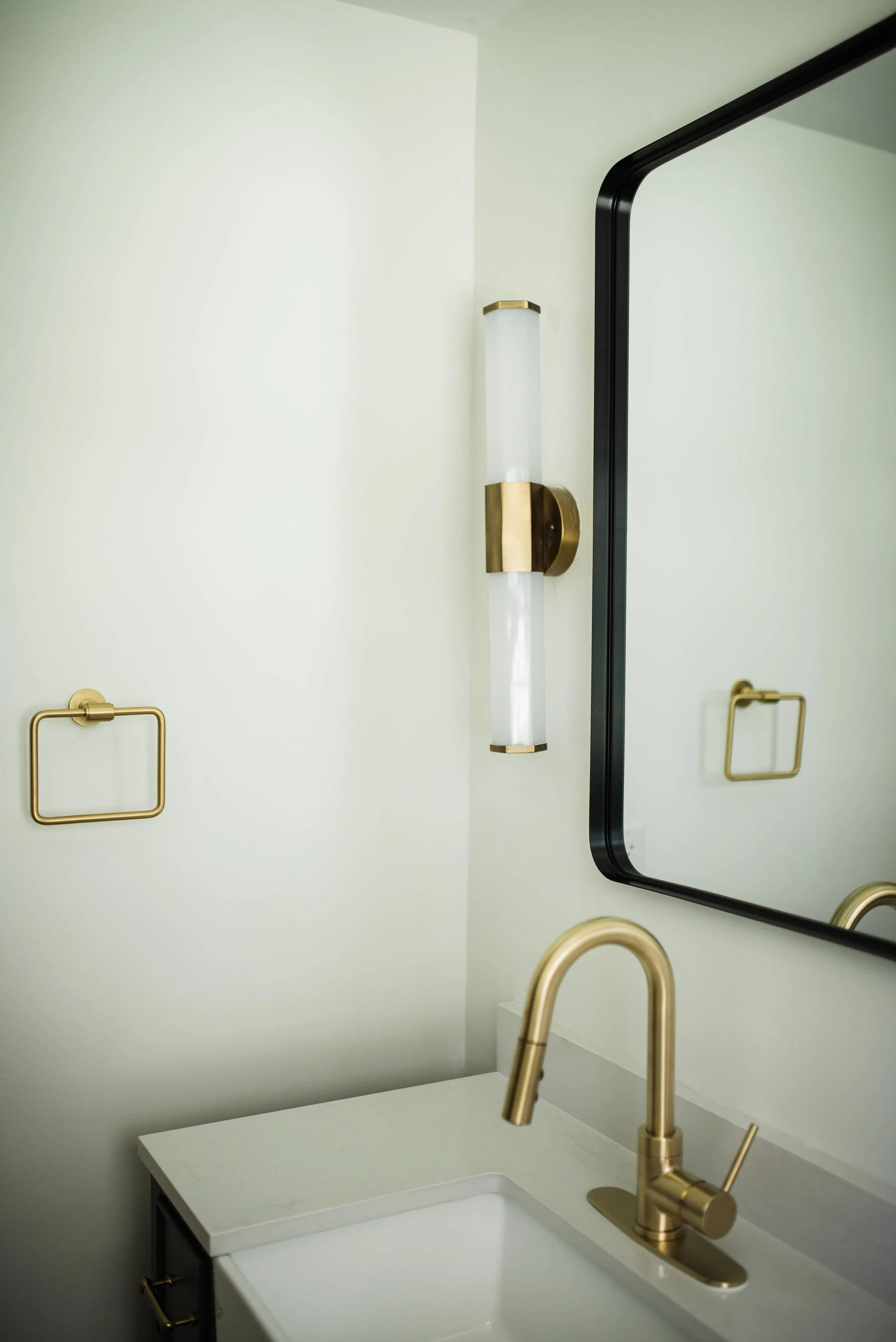 Modern bathroom with a black-framed mirror, gold fixtures including a faucet and towel holder, a white ceramic sink with a countertop, and a vertical wall light with gold accents.