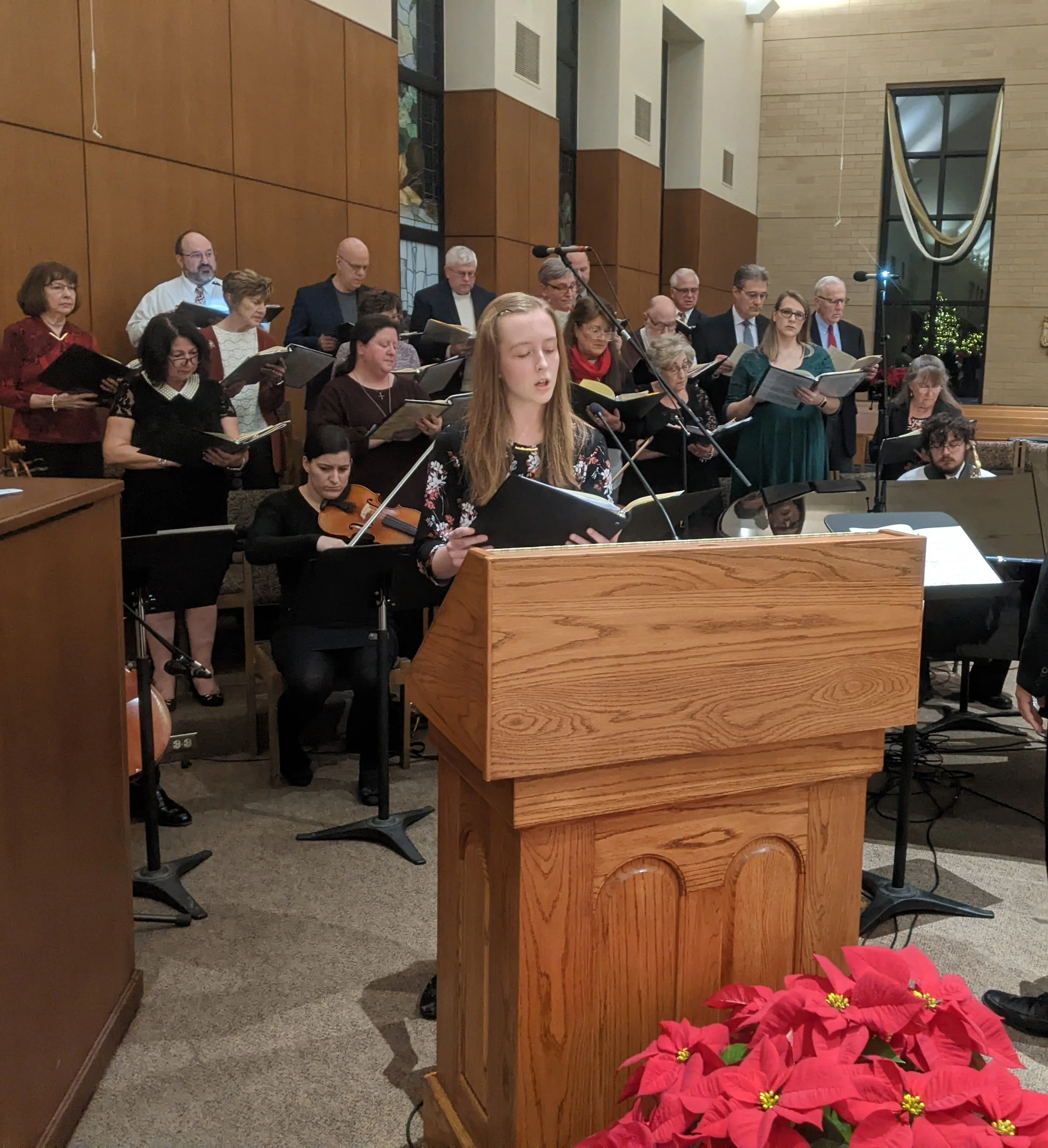 A choir of men and women singing and holding songbooks in a church with wooden walls and stained glass windows. A young woman is playing the violin in the foreground, and a poinsettia plant with red leaves is visible to the right.