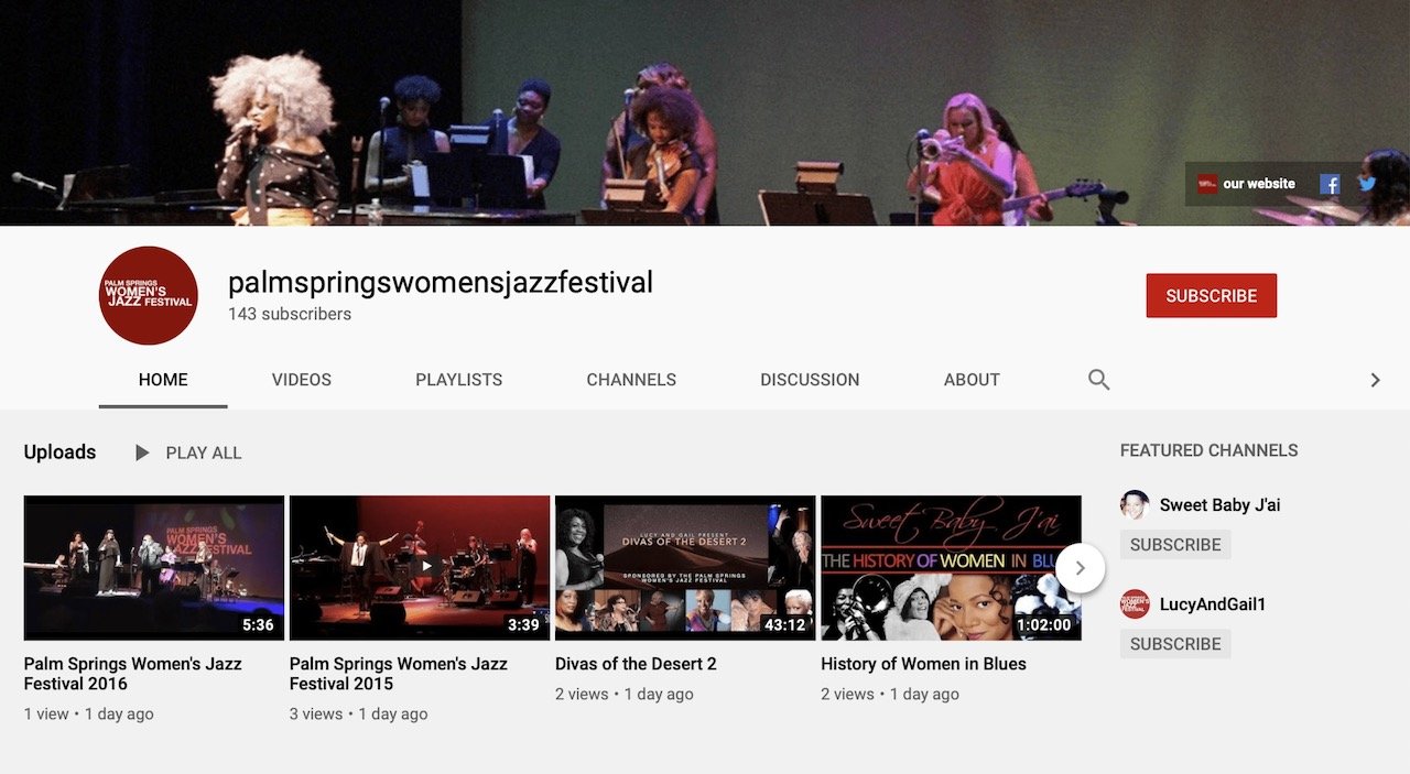 A stage with a group of musicians performing at the Palm Springs Women's Jazz Festival, including a woman with large curly hair singing, others playing instruments, and a backdrop displaying the festival's logo.