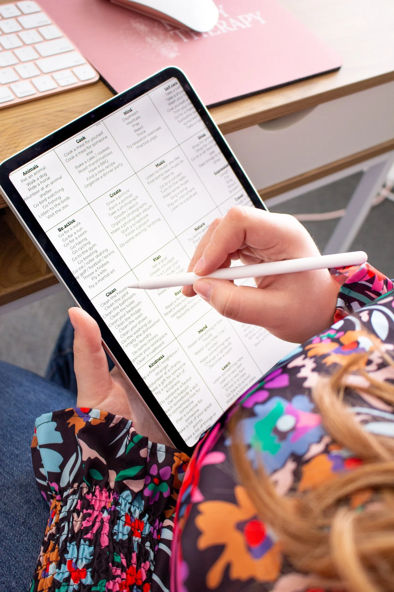 Person holding a tablet with a checklist, using a stylus to mark items, wearing a colorful floral jacket and sitting at a desk with a keyboard and notepad.