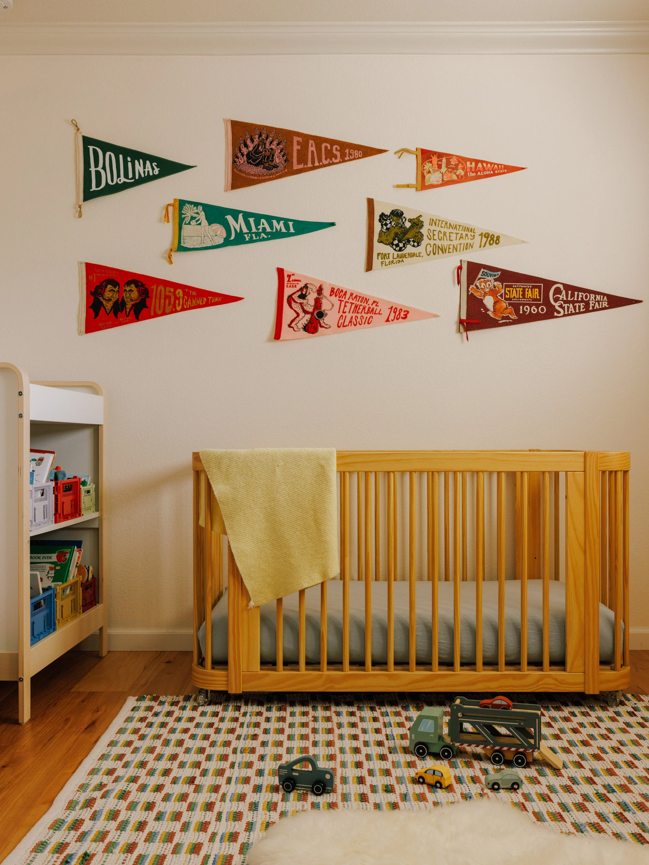 A child's bedroom designed by Mura Studio with a wooden bed, a colorful knitted rug, a blanket hanging on the side, a bookshelf on the left, and vintage-style souvenir flags hanging on the wall.