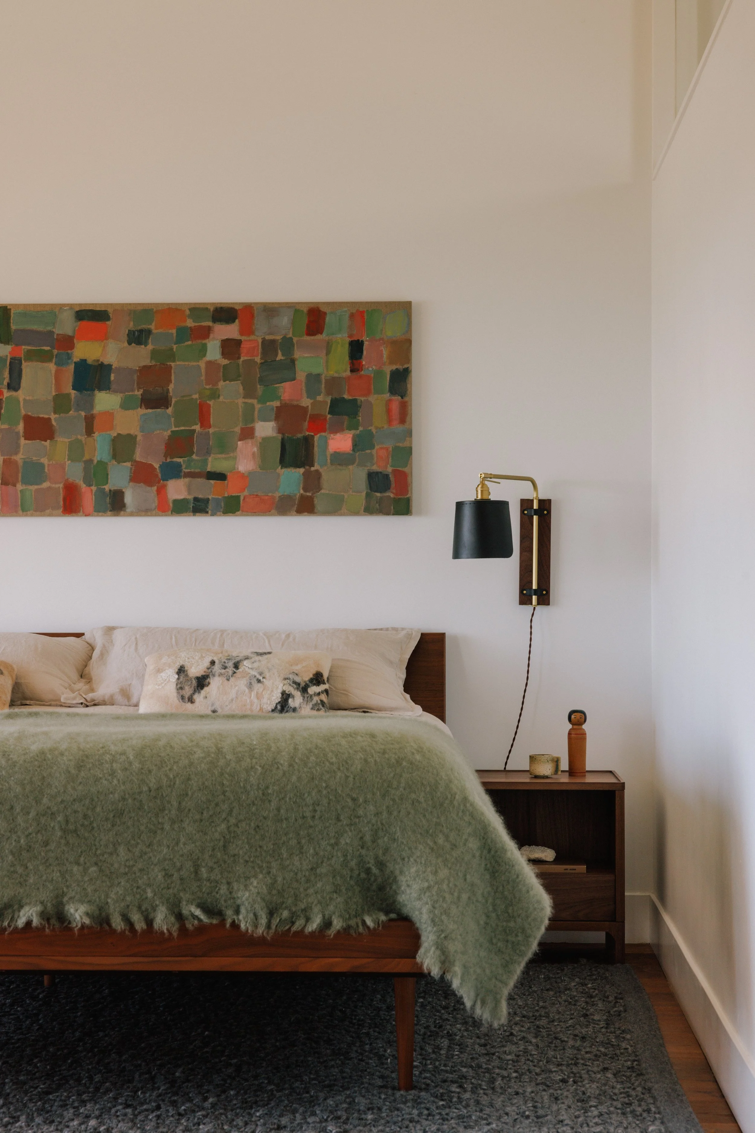 A cozy bedroom designed by Mura Studio with a wooden bed frame, a green fuzzy blanket, beige pillows, a small wooden nightstand with decorative items, a black and gold wall-mounted lamp, an abstract colorful painting, and a dark area rug.