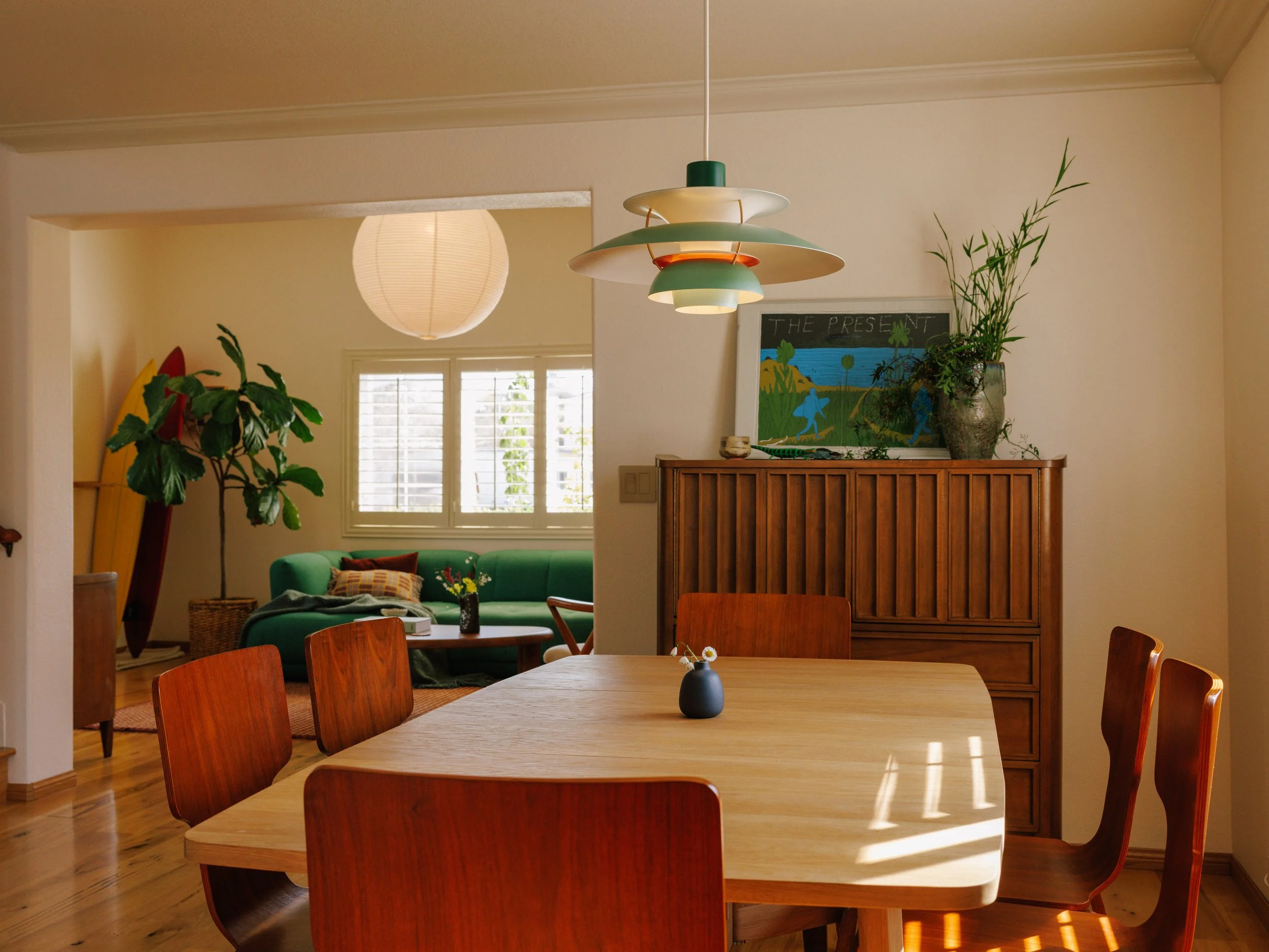 A cozy dining area with a wooden table and six matching chairs, a small vase with a flower, a wooden sideboard with plants and artwork on top, and a view into a living room with a green sofa, large window, and a paper lantern hanging from the ceiling.