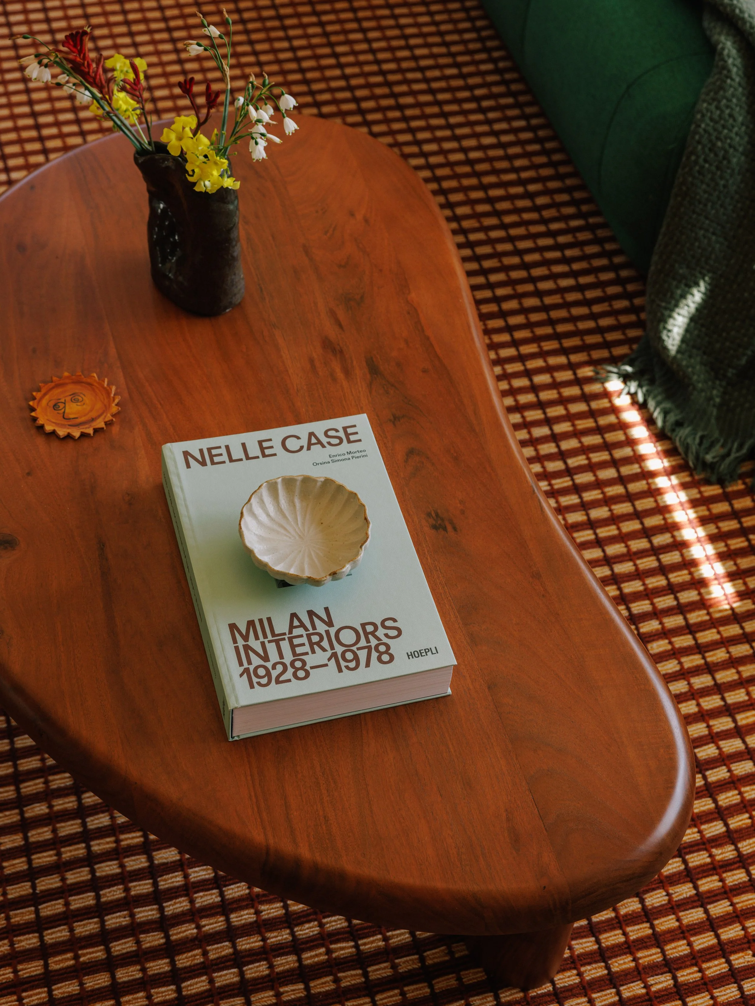 A wooden coffee table with a book, a small shallow bowl on top of the book, a dark ceramic vase with yellow and white flowers, a small orange decorative object with a face, and a green couch with a textured green blanket nearby. Styled by Mura Studio