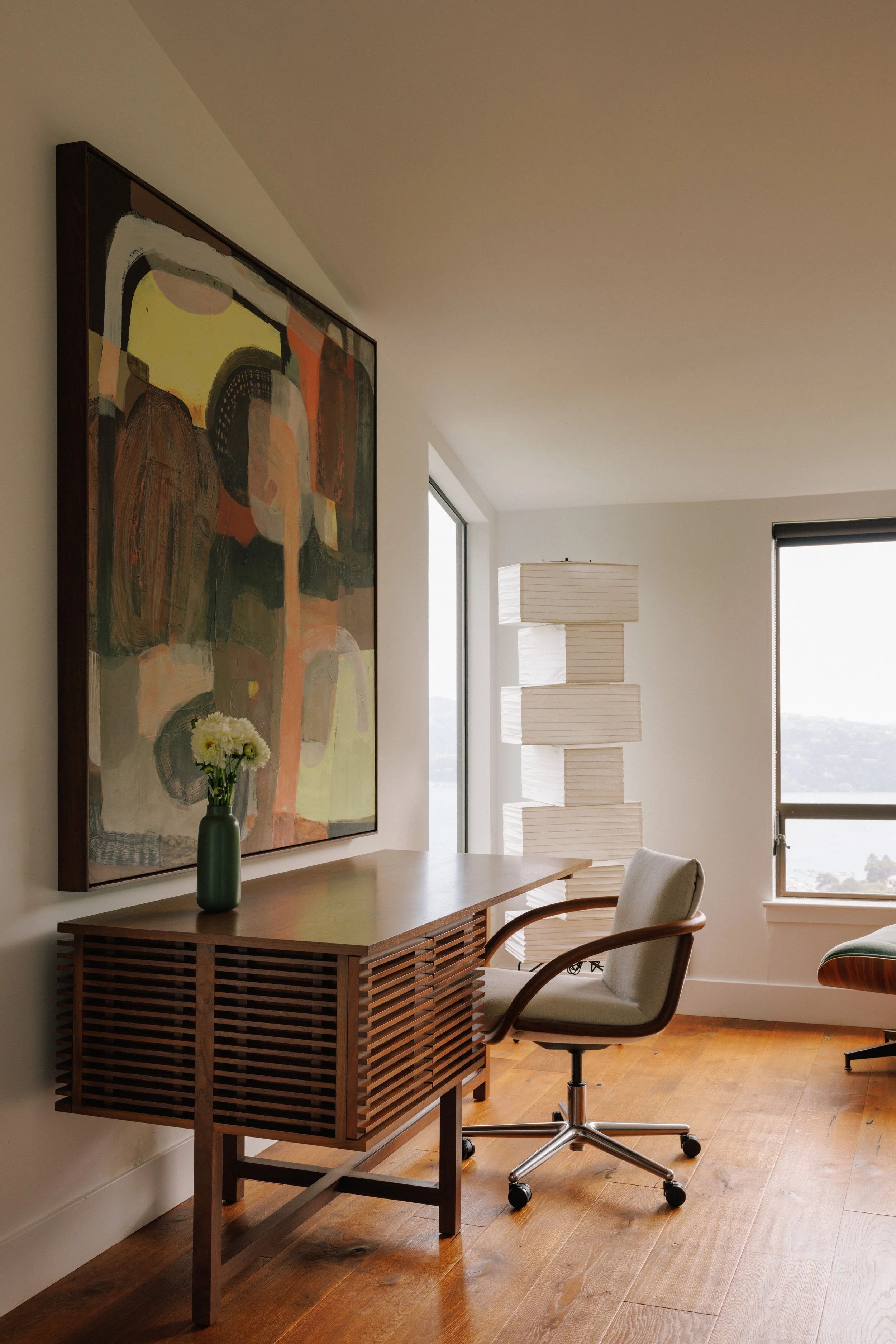 A home office designed by Mura Studio with a wooden desk, an beige office chair, a colorful abstract painting on the wall, a green vase with white flowers, large floor lamp with stacked rectangular shades, and two windows showing a view outside.