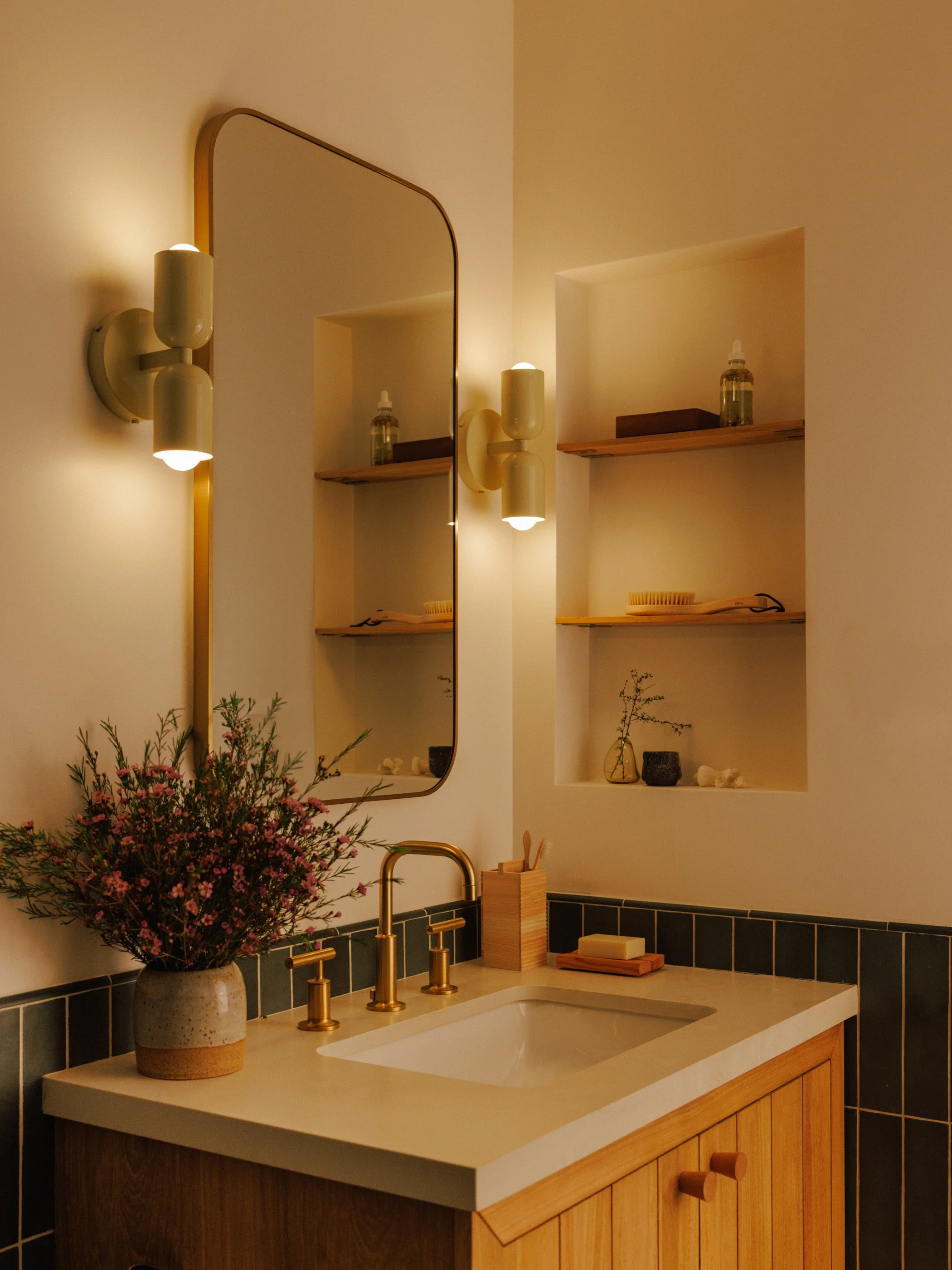 A bathroom vanity designed by Mura Studio with a white countertop, a rectangular sink, and a brass faucet. There is a mirrored cabinet above with two modern cream-colored wall sconces on either side.