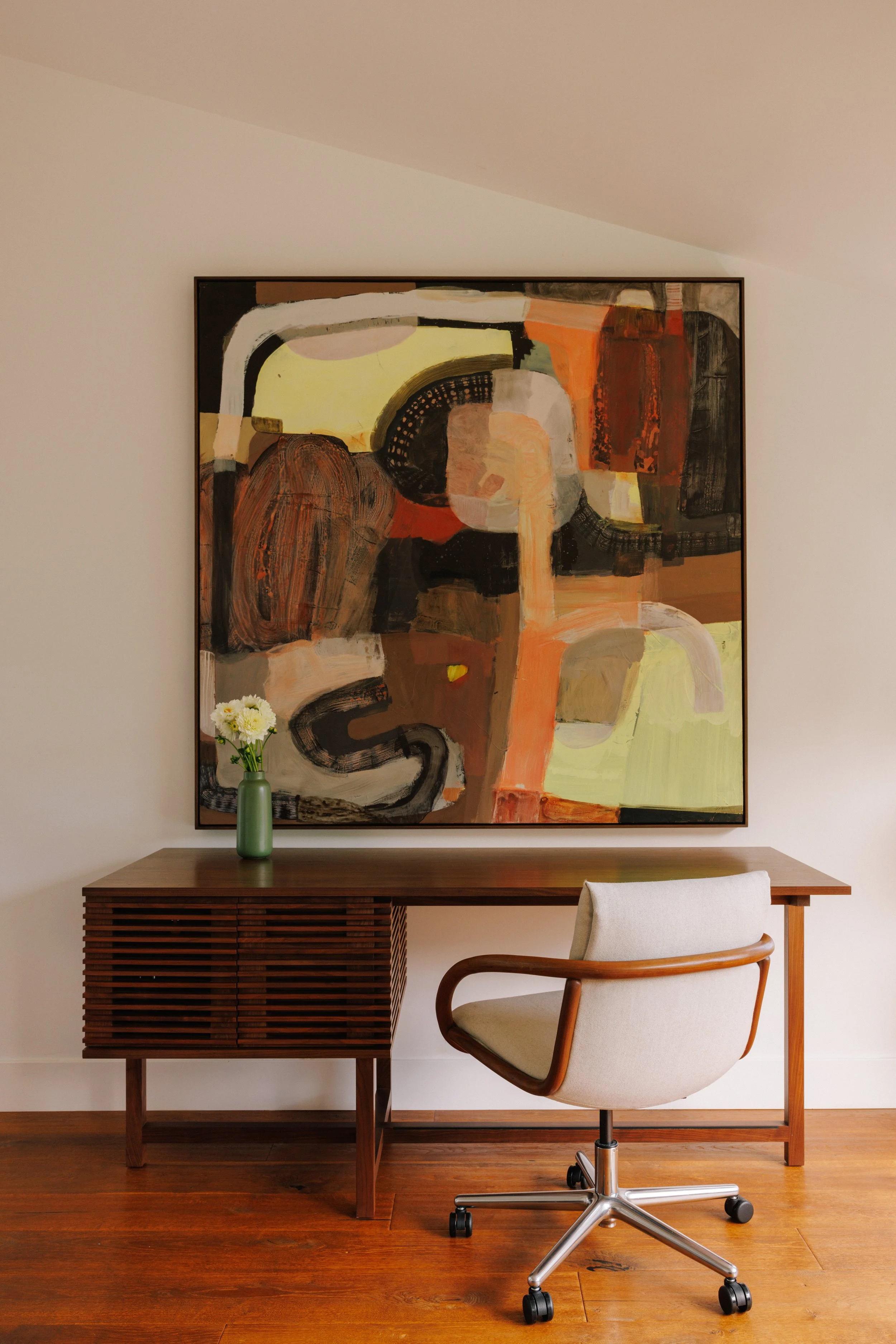 Minimalistic home office designed by Mura Studio with a wooden desk, an off-white office chair, a large abstract painting on the wall, and a green vase with white flowers on the desk.