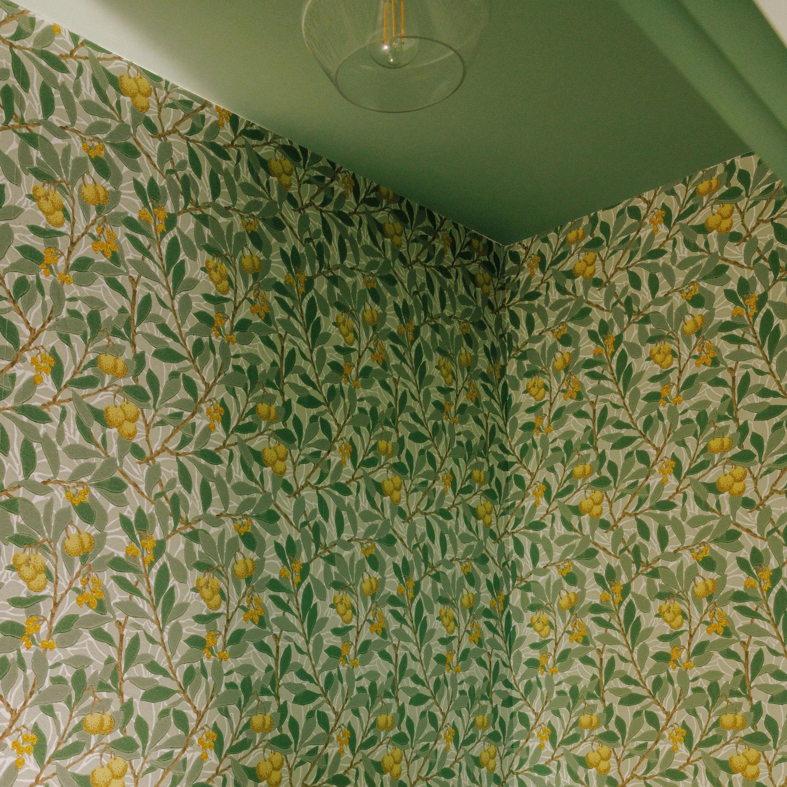 A ceiling light fixture hangs above a wall with floral wallpaper featuring green leaves and yellow berries, designed by Mura Studio.