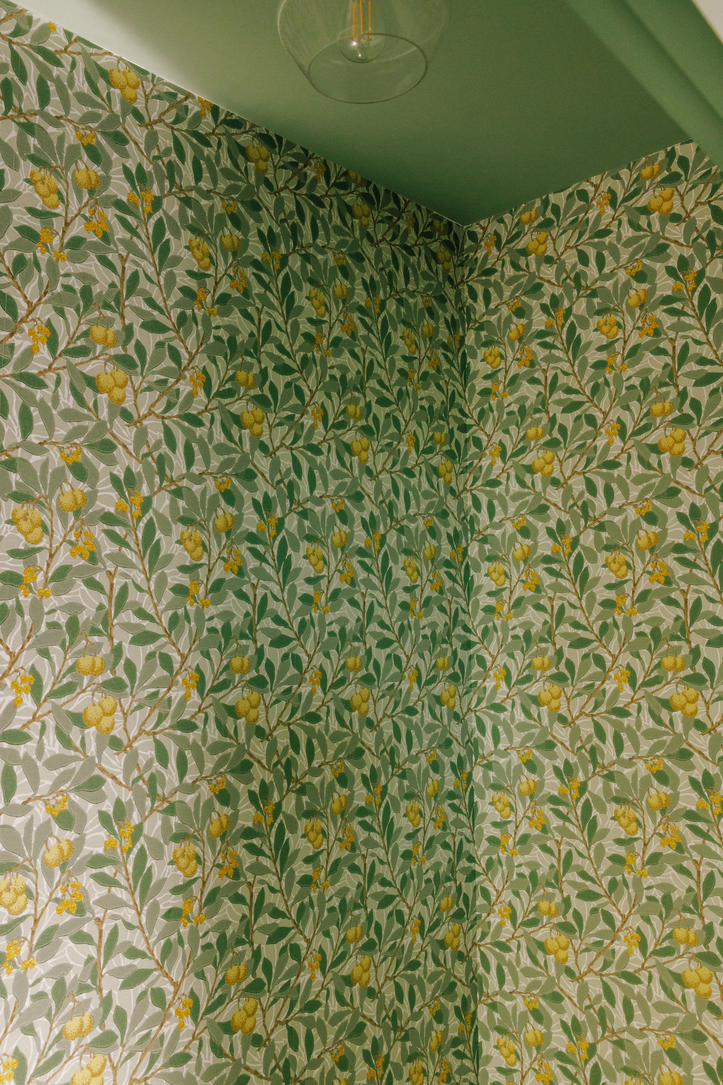 Wallpaper with green leaves and yellow fruit pattern on walls, with a glass ceiling light fixture visible at the top, interior design by Mura Studio.