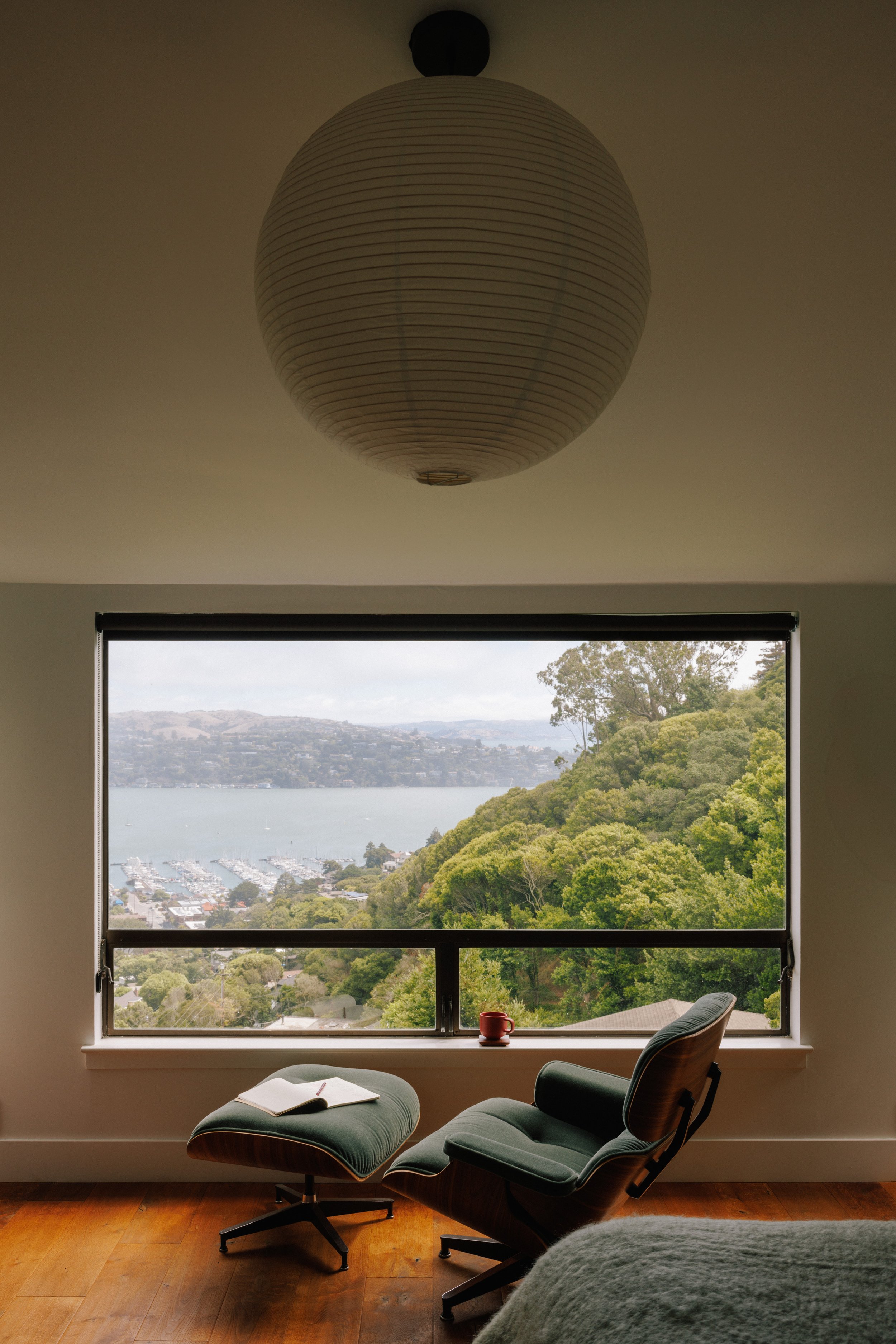 Room designed by Mura Studio with large window view of green hills, water, and marina. Interior has lounge chair with matching footrest and open book, red mug on windowsill, and a round paper lantern ceiling light.