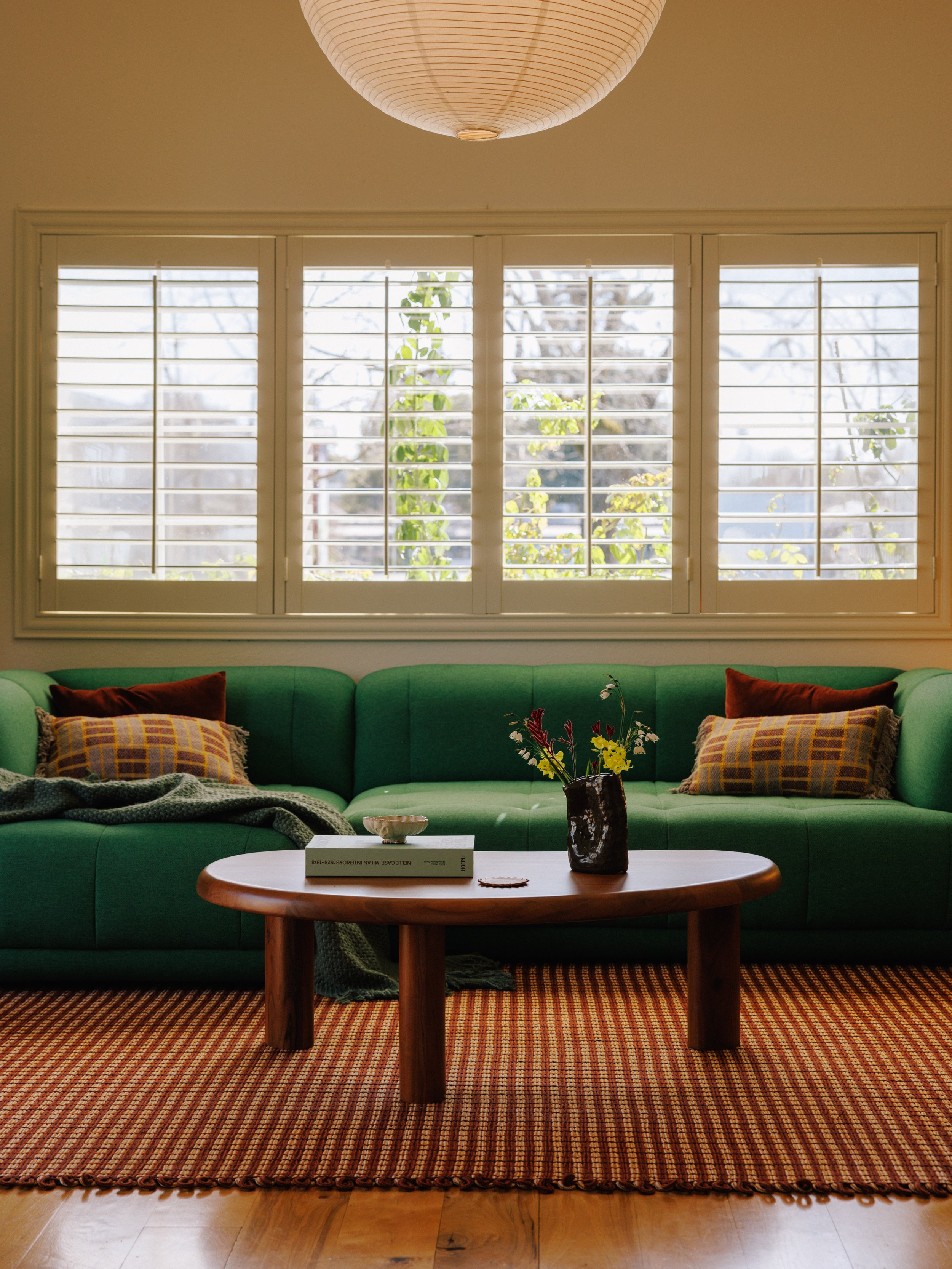 Living room by Mura Studio with a green sofa, two plaid pillows, a blanket, a coffee table with a book and a vase of flowers, large windows with shutters, and a hanging paper lantern.