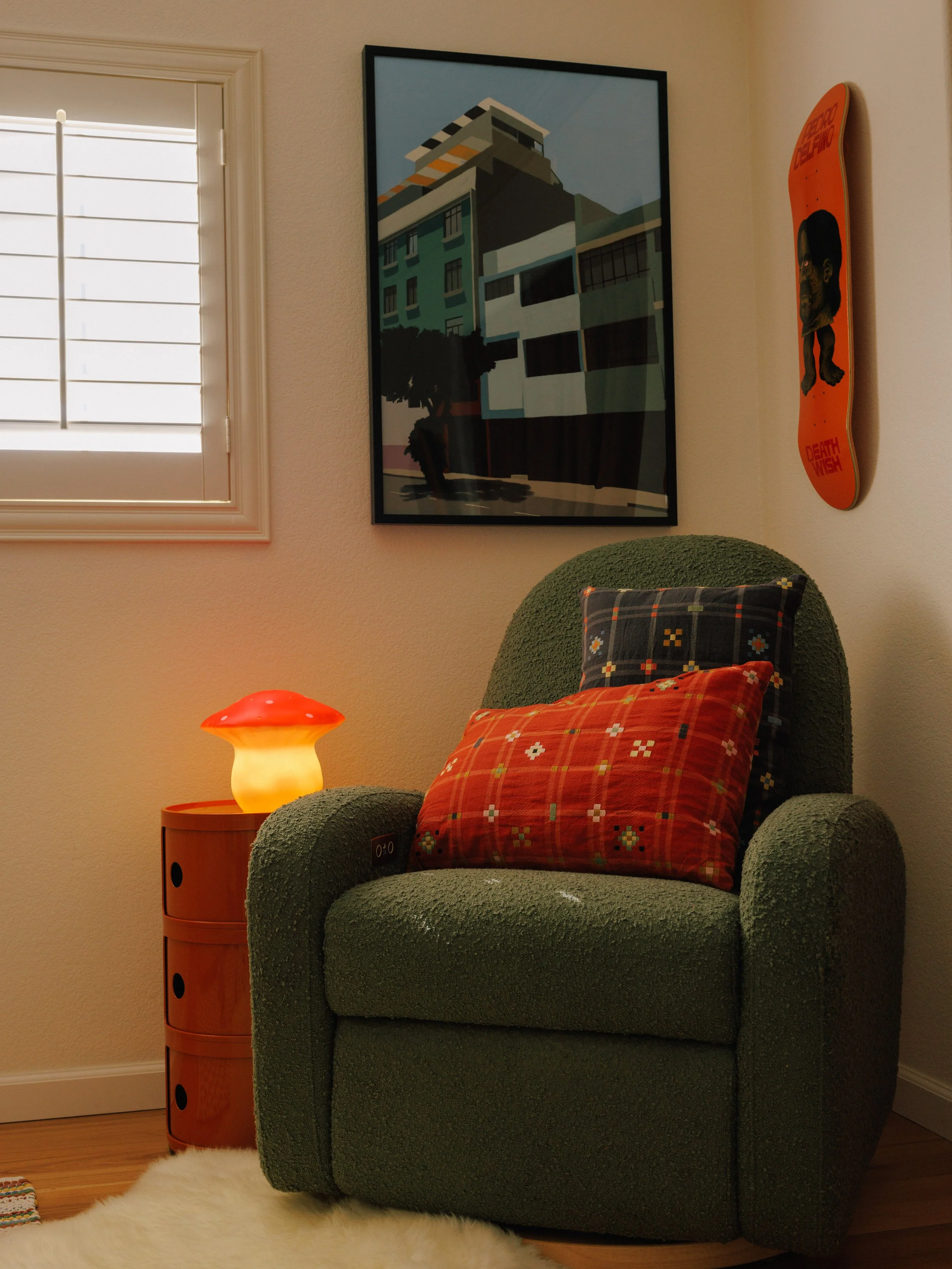 A cozy corner with a green armchair, two patterned throw pillows, a small table with a lamp, a window with white blinds, a large framed picture of a modern building on the wall, and a skateboard on the wall, interior design by Mura Studio.