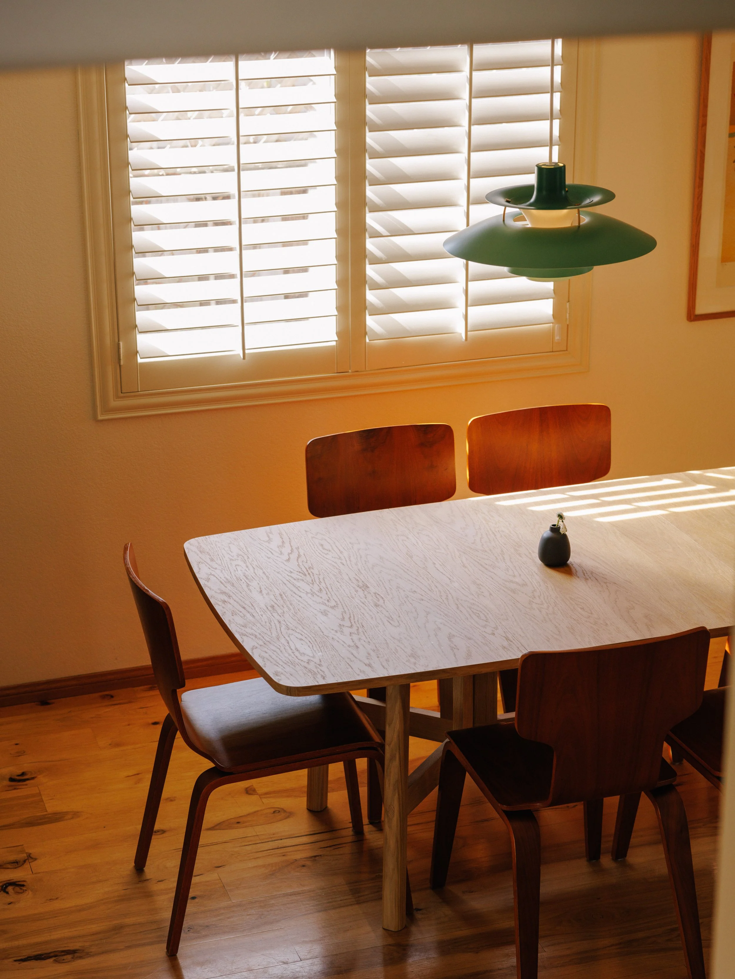 A dining room designed by Mura Studio with a light wood table, four matching wooden chairs, a vase in the center, and a window with white plantation shutters. A green pendant light hangs above the table.