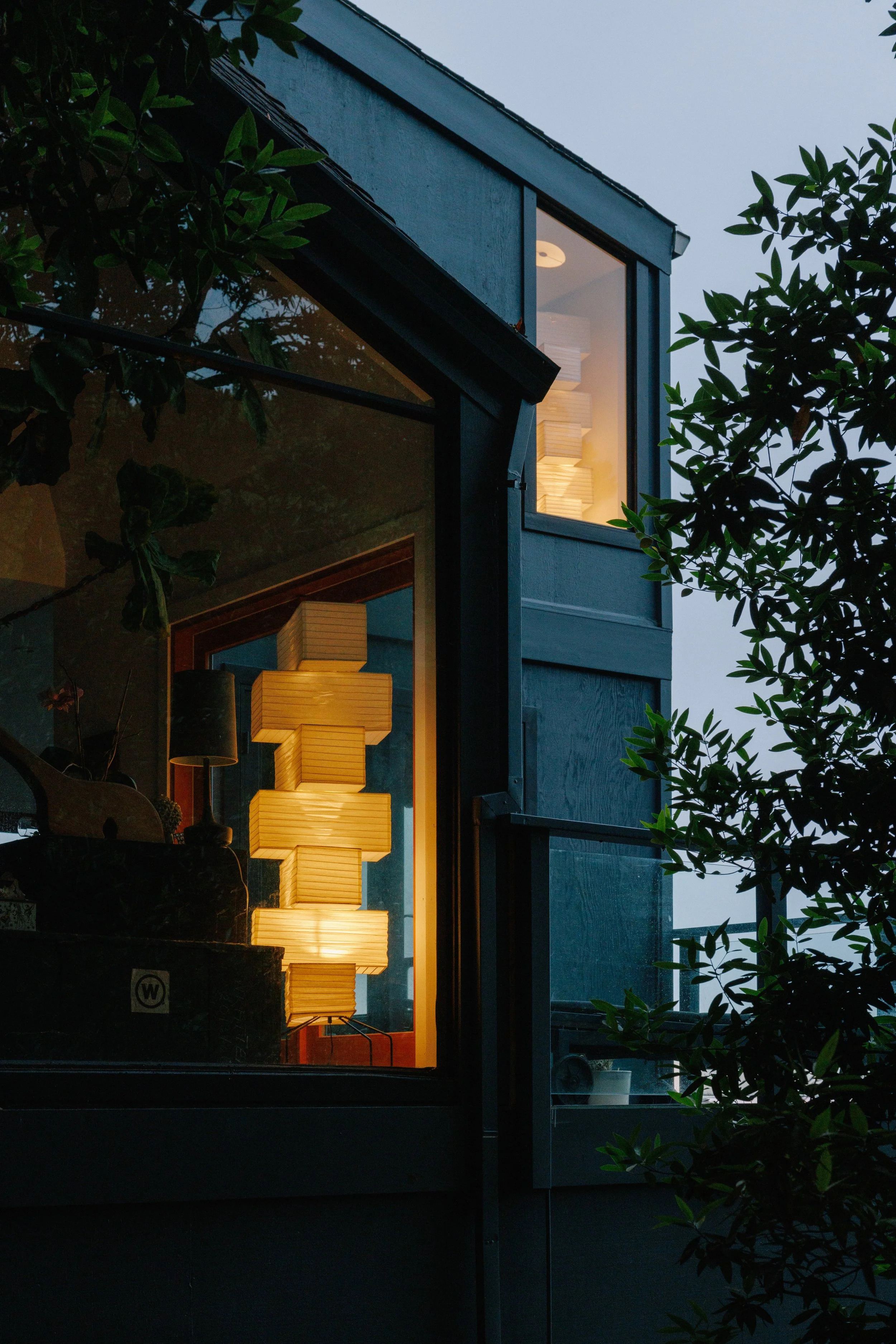 A modern house with interior design by Mura Studio with large glass windows; illuminated interior shows a tall, stacked lamp and other furnishings; exterior is dark with trees partially blocking view.