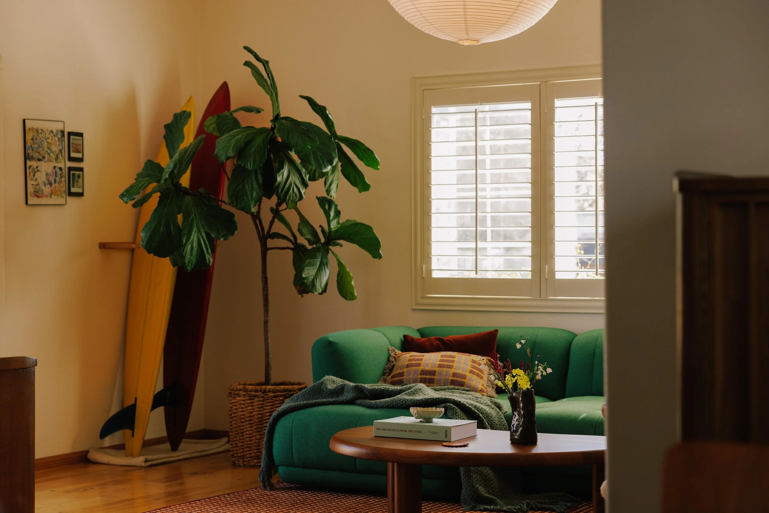 Cozy living room designed by Mura Studio with a green sofa, a window with white blinds, and a potted plant.