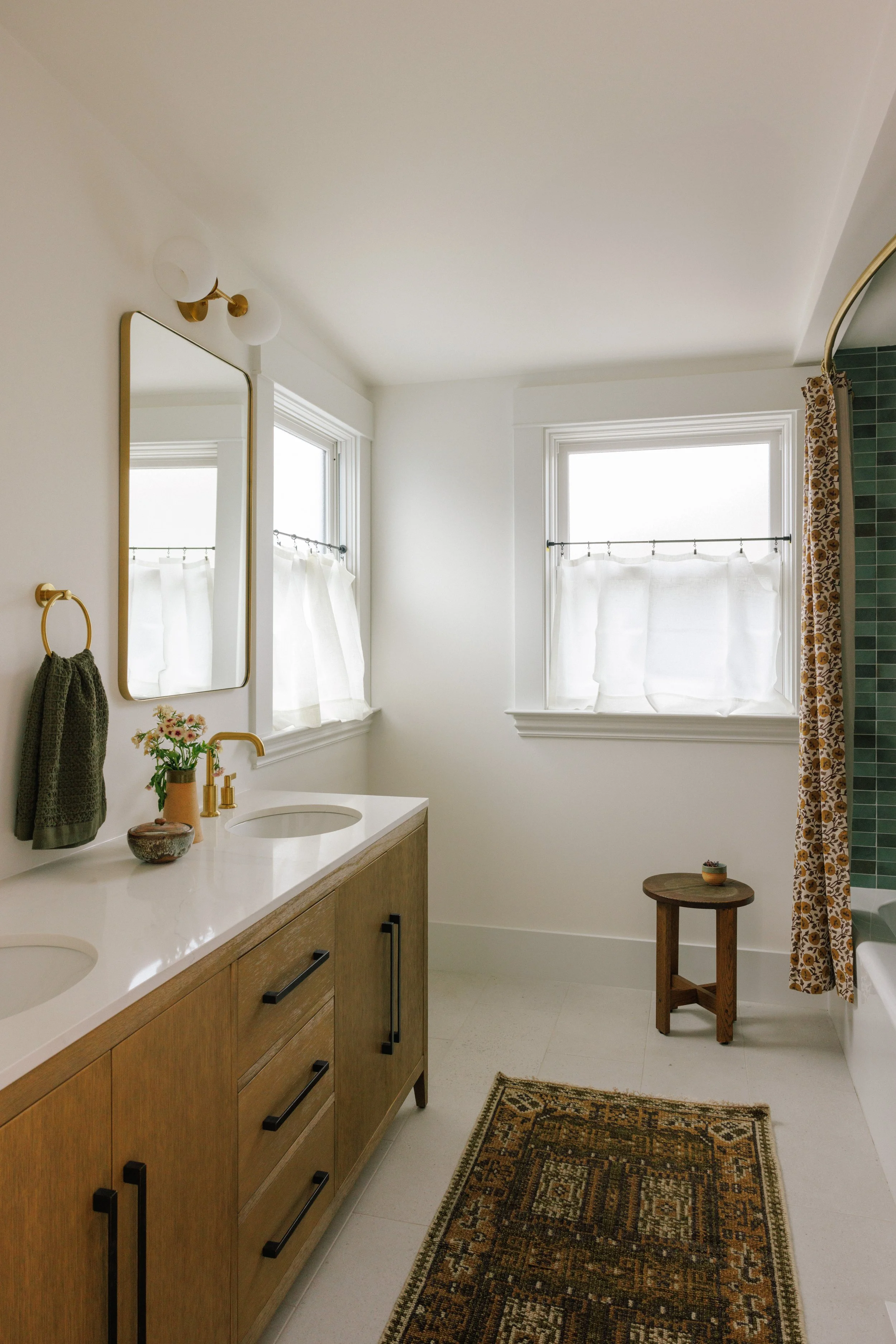 Modern bathroom designed by Mura Studio with white walls, a double wooden vanity with white countertop, gold fixtures, and a mirror. Two windows with white curtains, a small wooden stool, a patterned curtain to the bathtub, and a decorative rug.