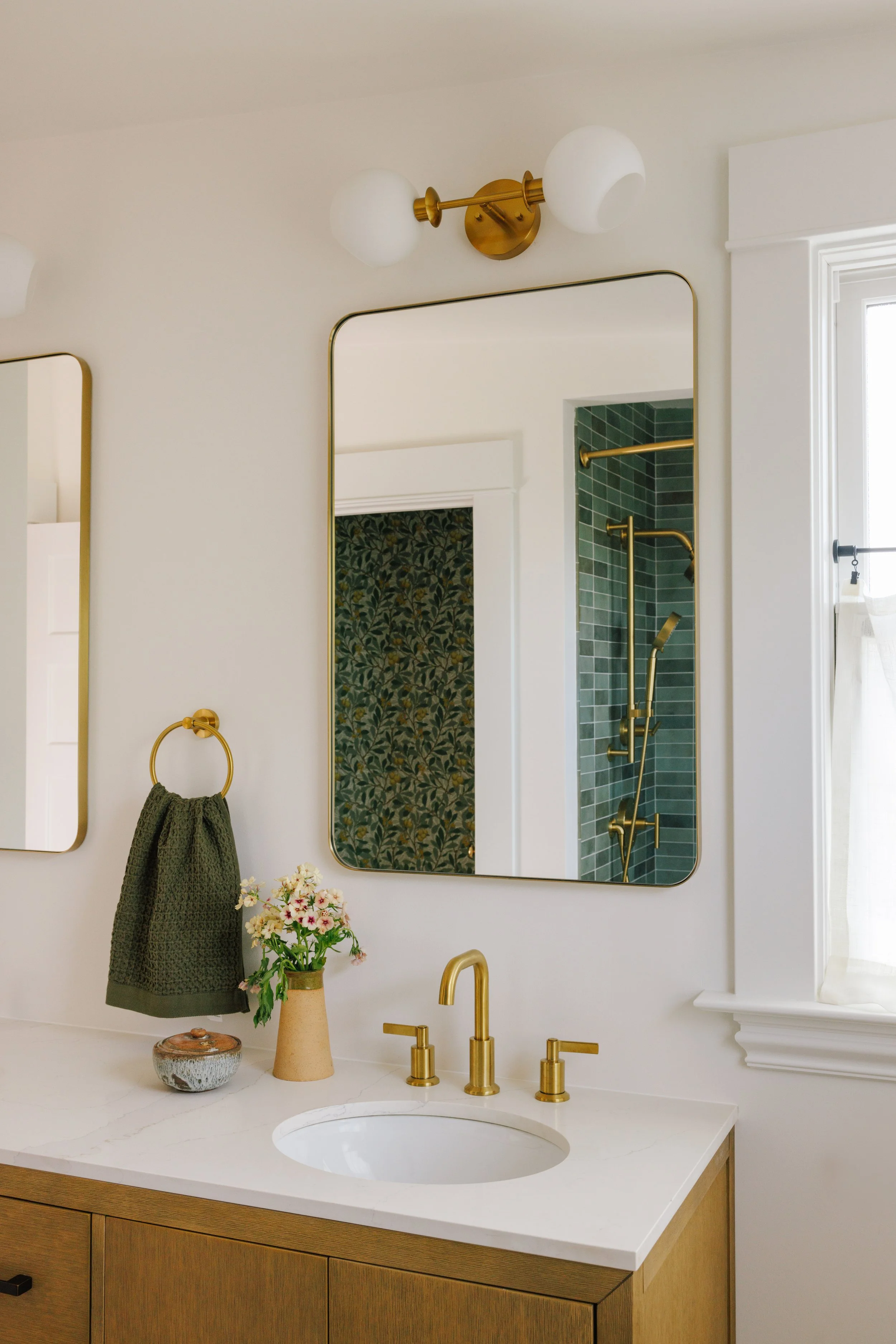 A bathroom vanity designed by Mura Studio with a white countertop, gold fixtures, a vase of flowers, and a mirror. In the reflection, a doorway with patterned wallpaper and a shower with green tiles and gold hardware are visible.