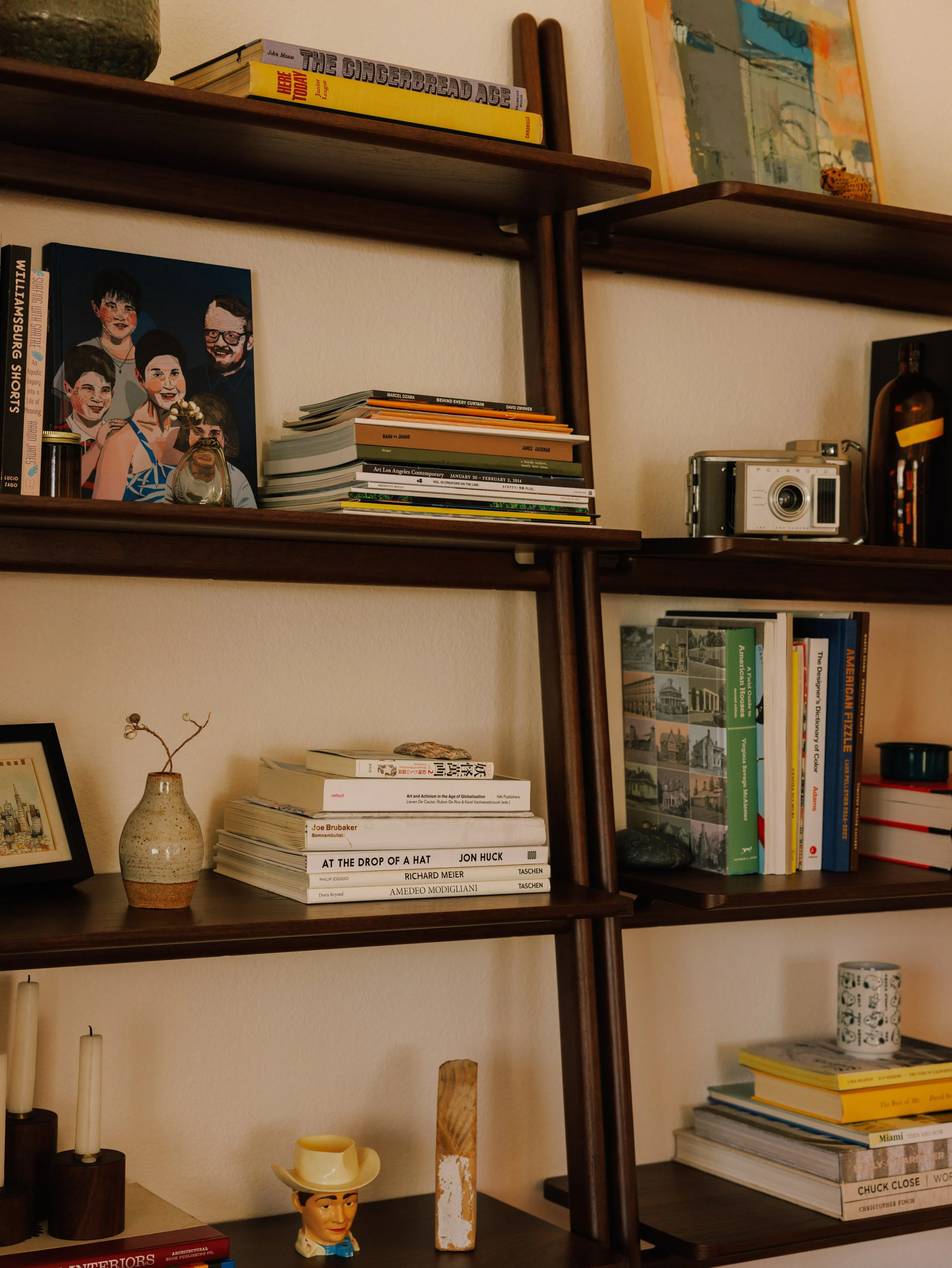 Wooden wall shelves with books, magazines, and decorative items including a ceramic vase, a vintage camera, and artwork styled by Mura Studio.
