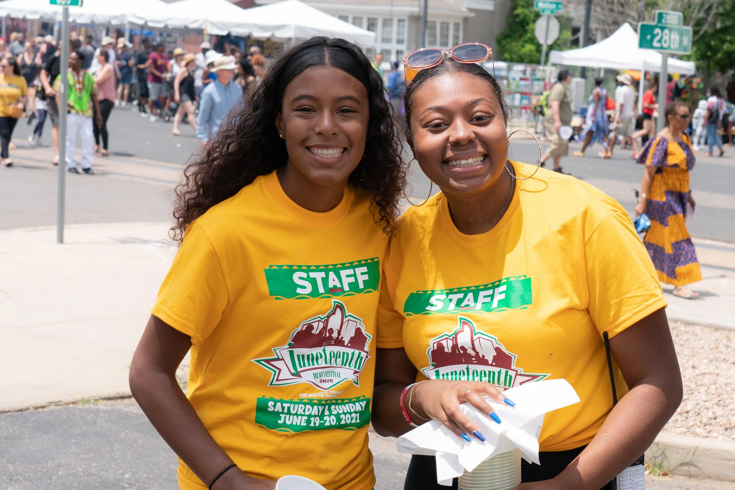 Two women smiling outdoors at a festival, wearing yellow T-shirts with the text "Luncheon" and the event details, with a crowded street fair background.