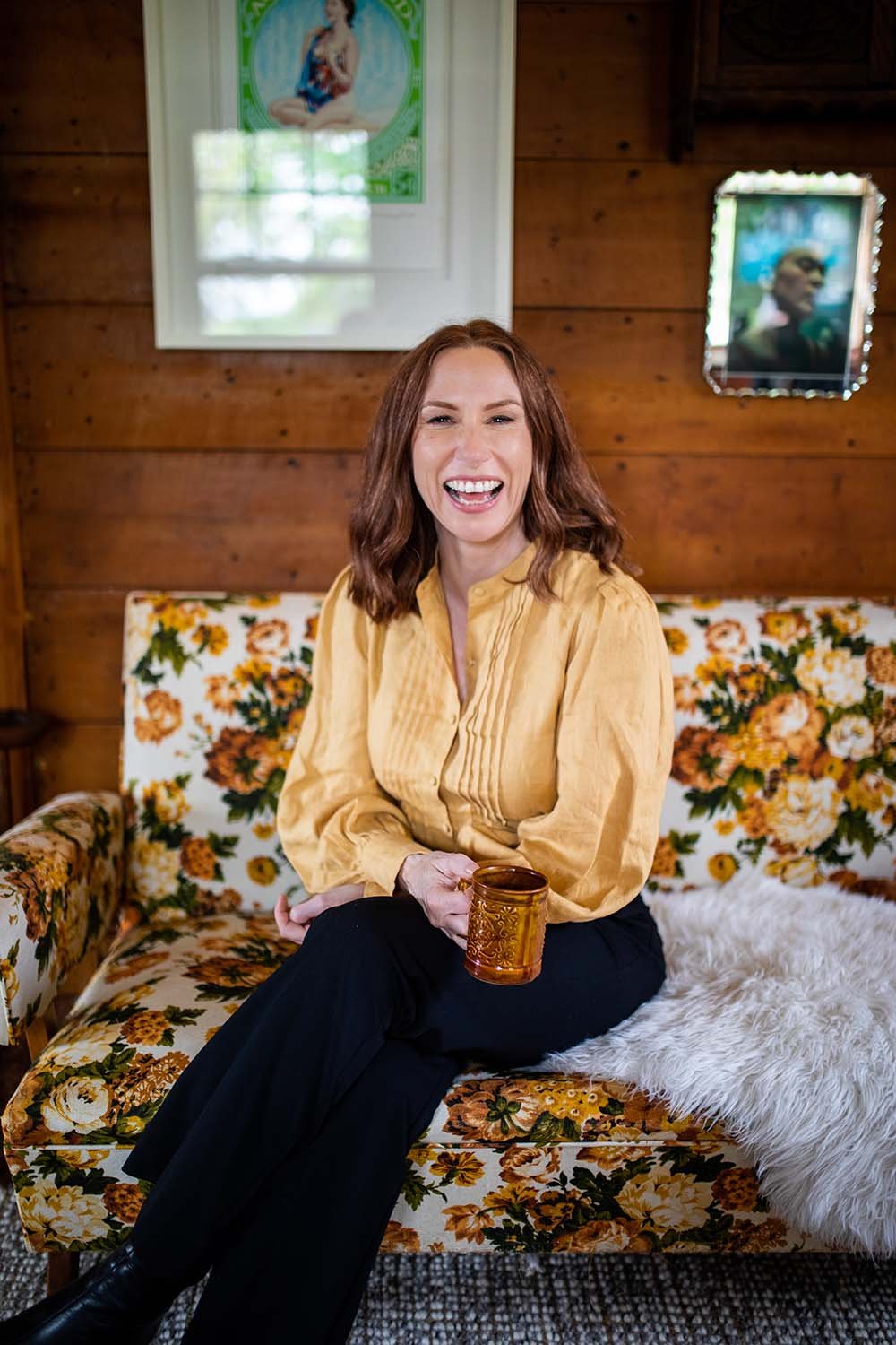 Woman sitting on floral couch in yellow shirt laughing with cup of tea in hand