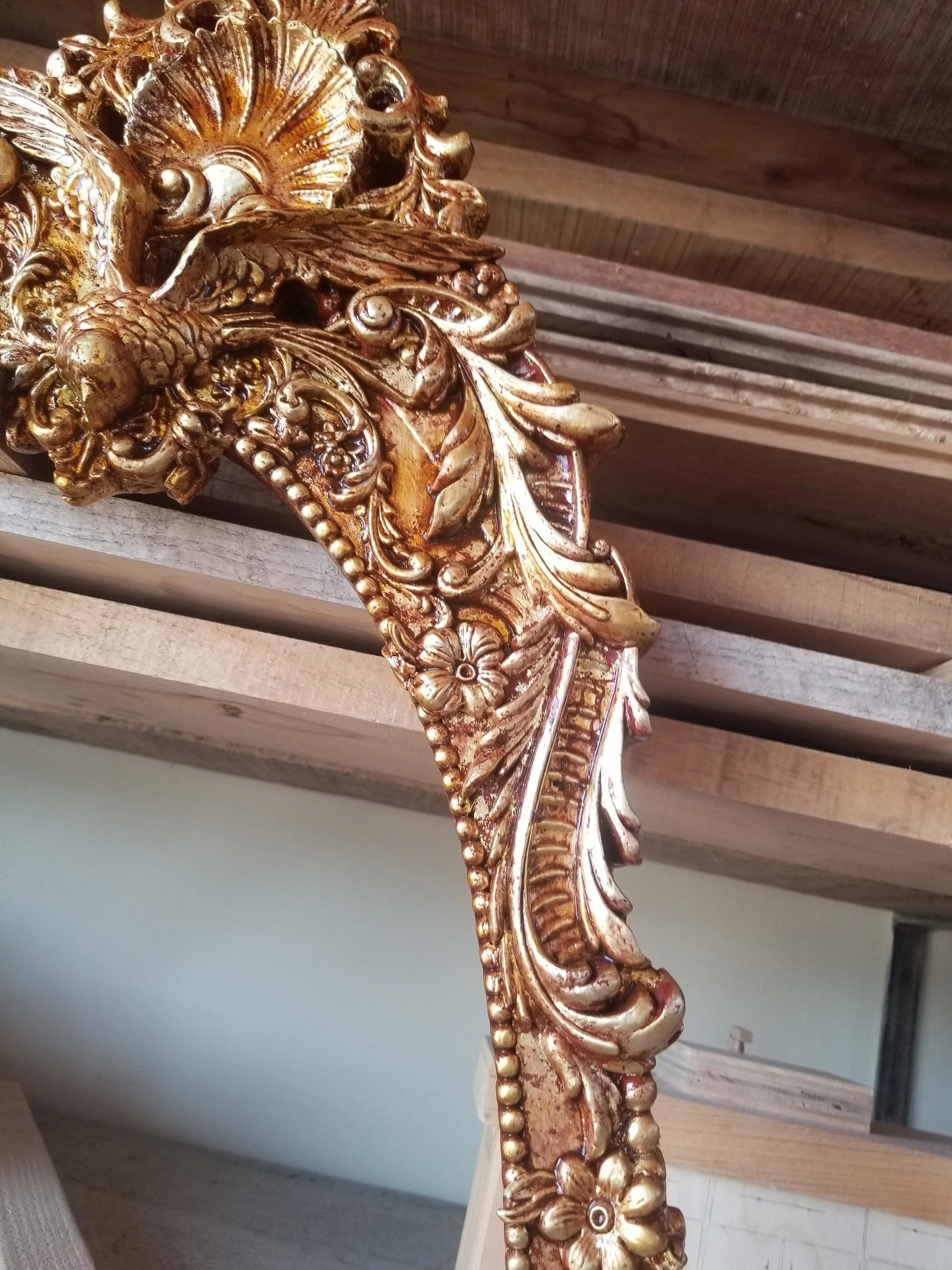 Close-up of an ornate, carved, gold-colored decorative frame with floral and leaf patterns, resting on a wooden surface.