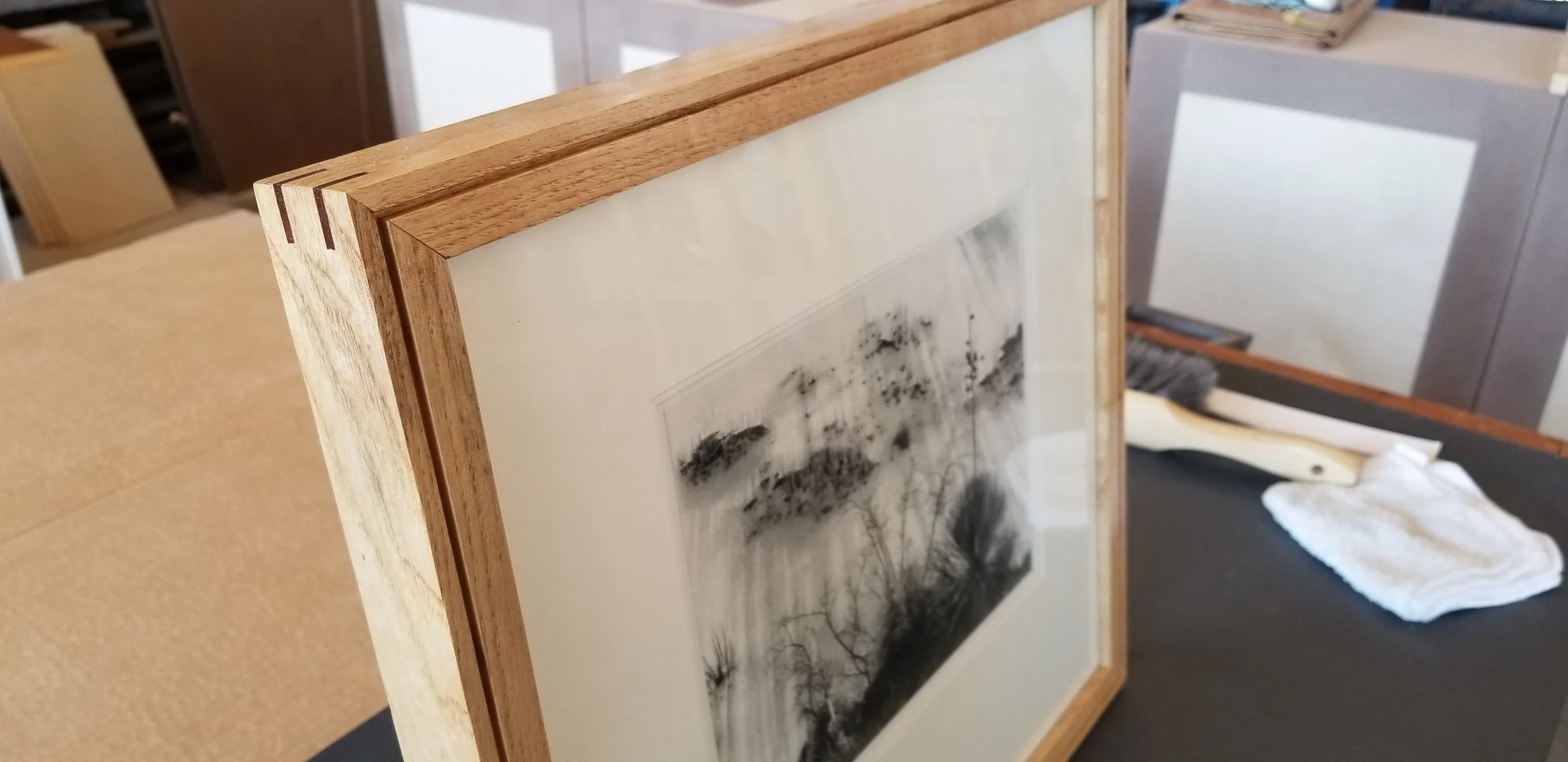 Framed black-and-white landscape photograph featuring trees, mounted on a table with cleaning cloth and squeegee nearby.