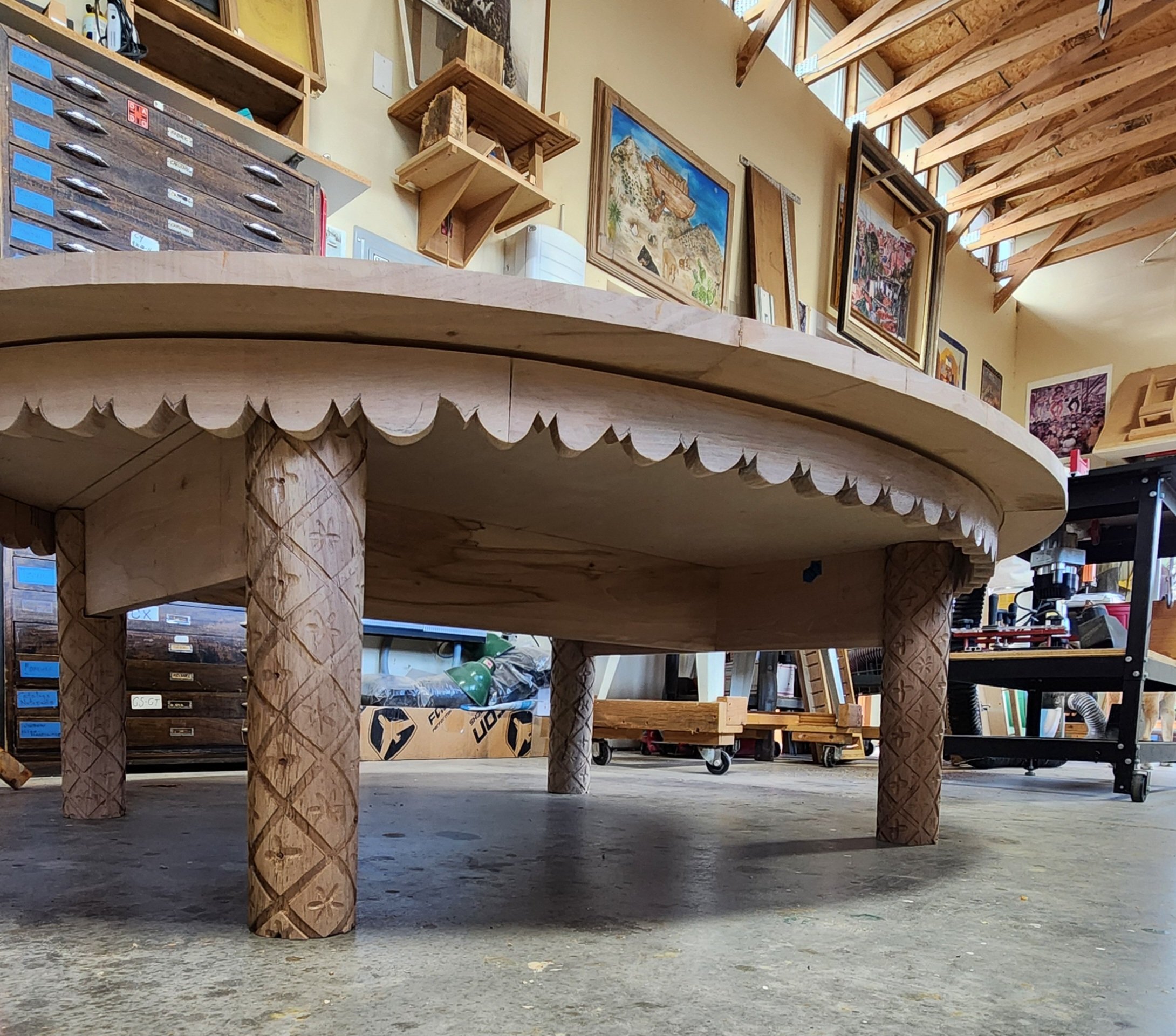 A wooden table with intricately carved legs and a scalloped edge in a woodworking shop, with framed artwork on the wall above and various tools and supplies in the background.