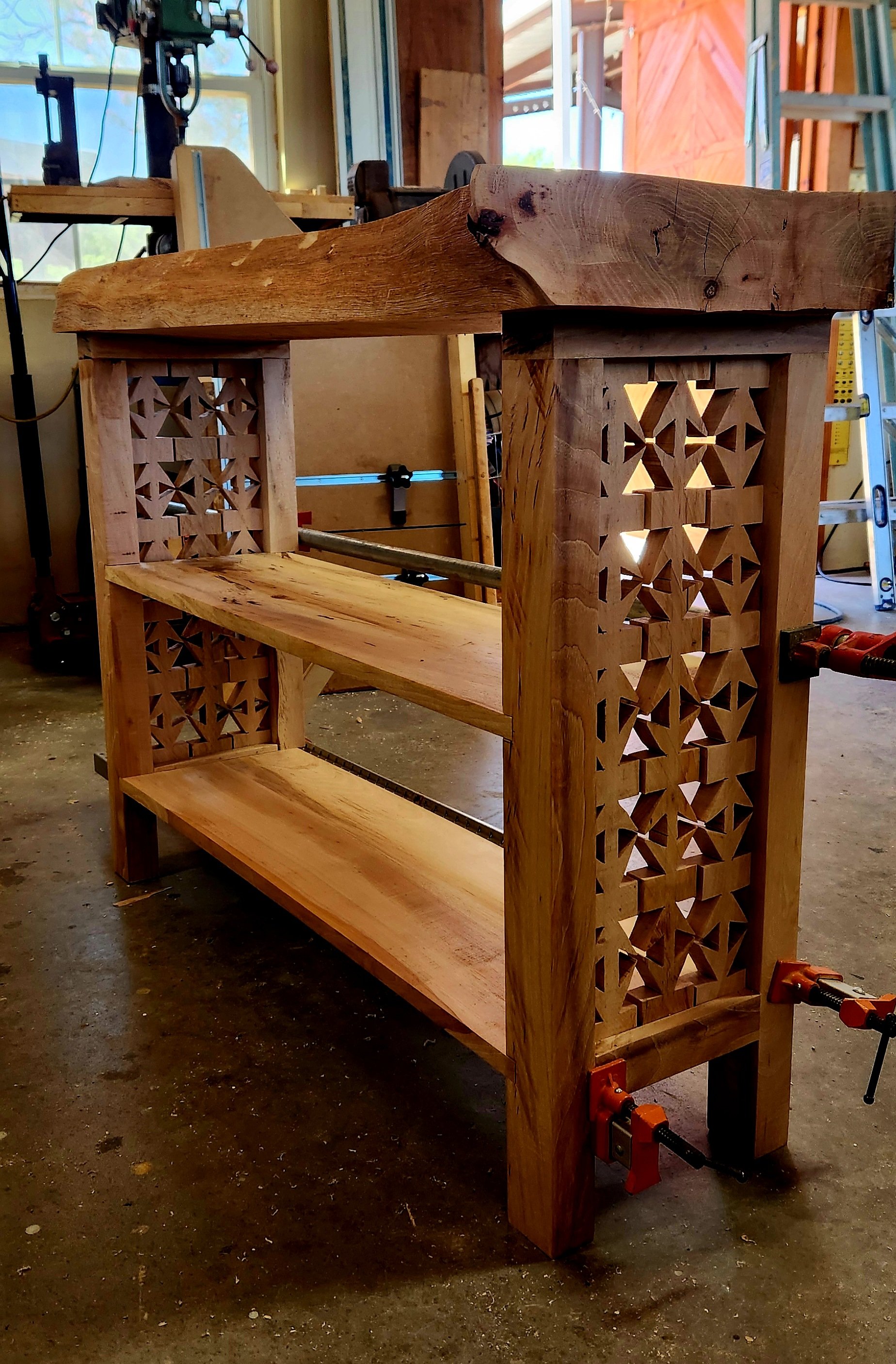 A wooden shelving unit with decorative carved sides in a woodworking workshop, clamps holding parts in place, and tools and construction materials around.