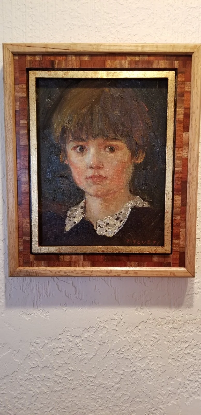 A portrait painting of a young girl with short light brown hair, wearing a white lace collar, set in a wooden frame on a white textured wall.