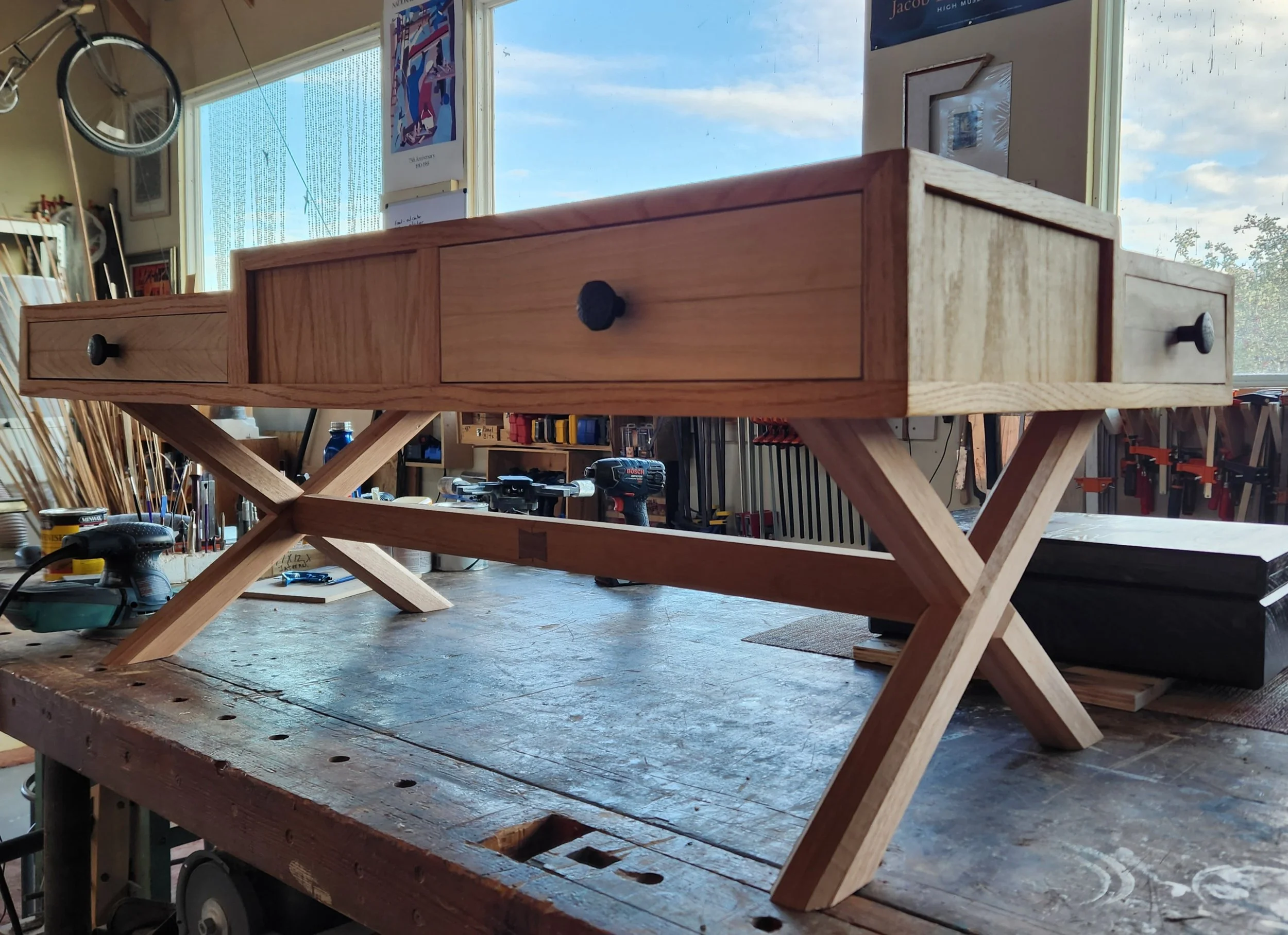 A wooden table with drawers and a crisscross wooden base in a woodworking workshop, surrounded by tools and materials, with a window showing blue sky and clouds outside.