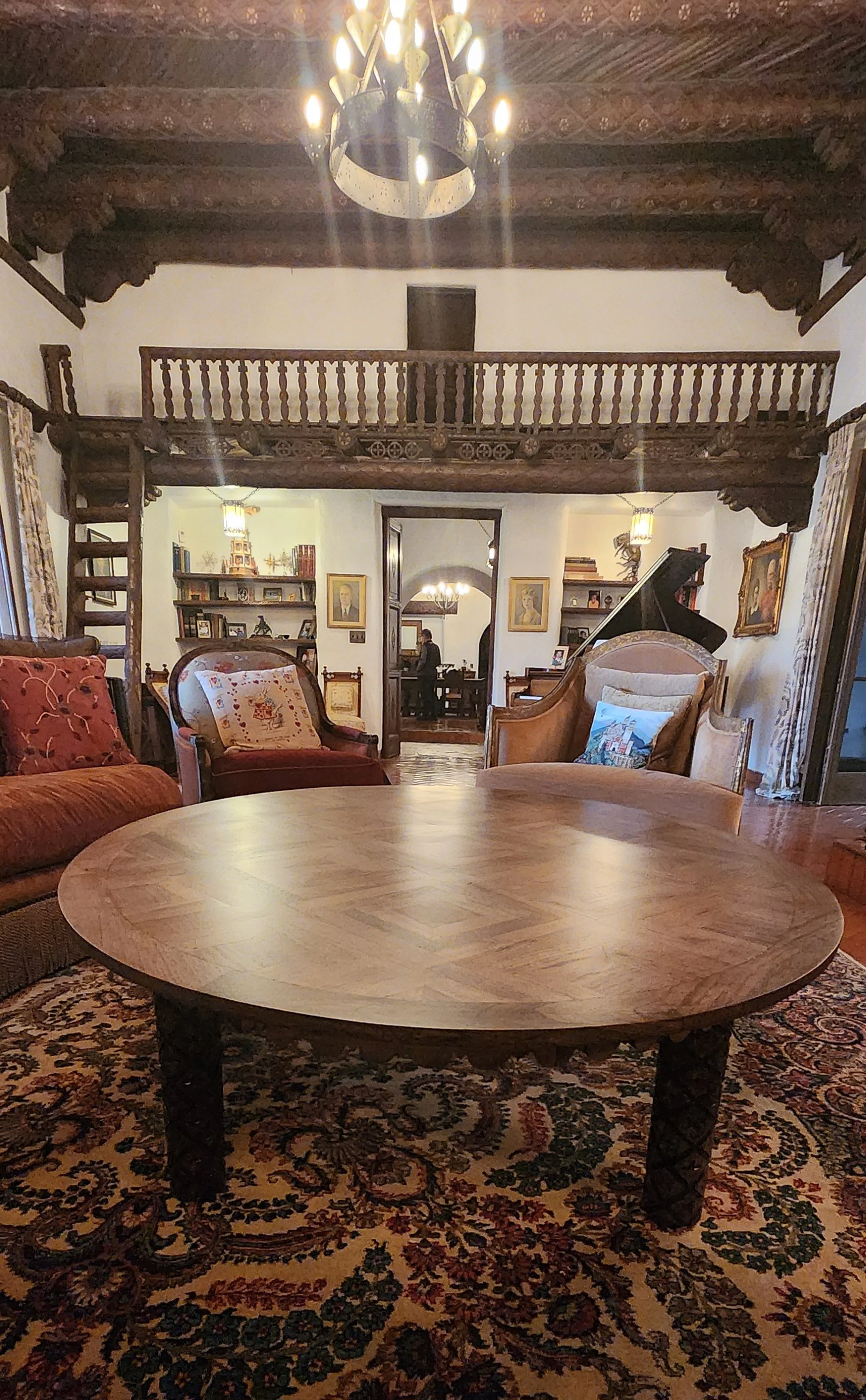 A cozy, vintage-style living room with a large round wooden coffee table, upholstered armchairs with decorative pillows, a patterned area rug, and a grand piano. The room features wooden beams on the ceiling, a chandelier, bookshelves, framed portrai