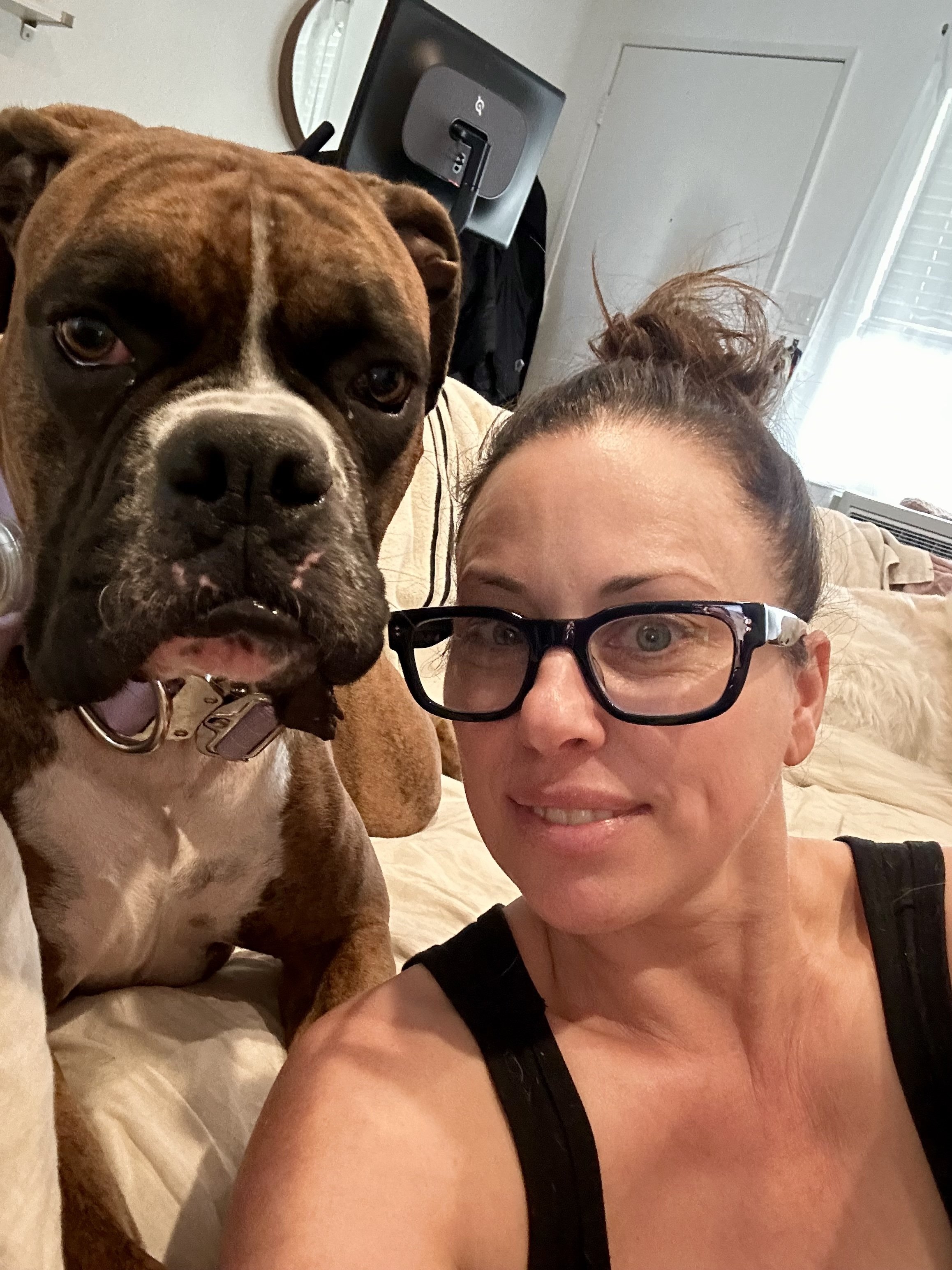A woman with glasses and a black tank top taking a selfie with a large brown and white Boxer breed, in a cozy living room.