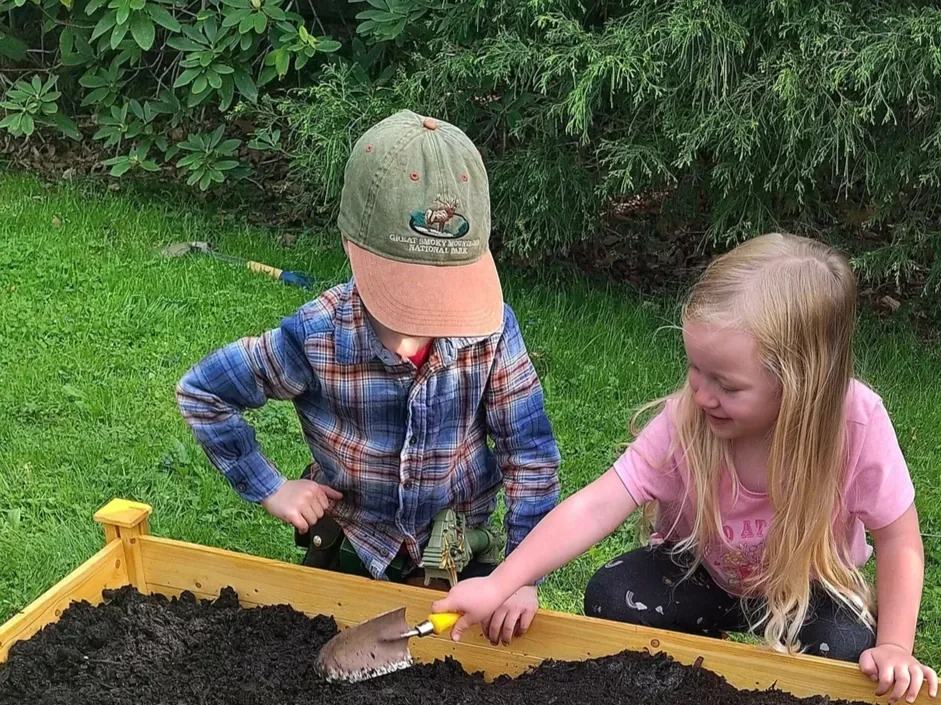 A boy and girl are playing in a garden with a wooden sandbox filled with dark soil. The boy is wearing a green cap and plaid shirt, while the girl is wearing a pink shirt and has long blonde hair. They are smiling and appear to be enjoying digging in the soil together.