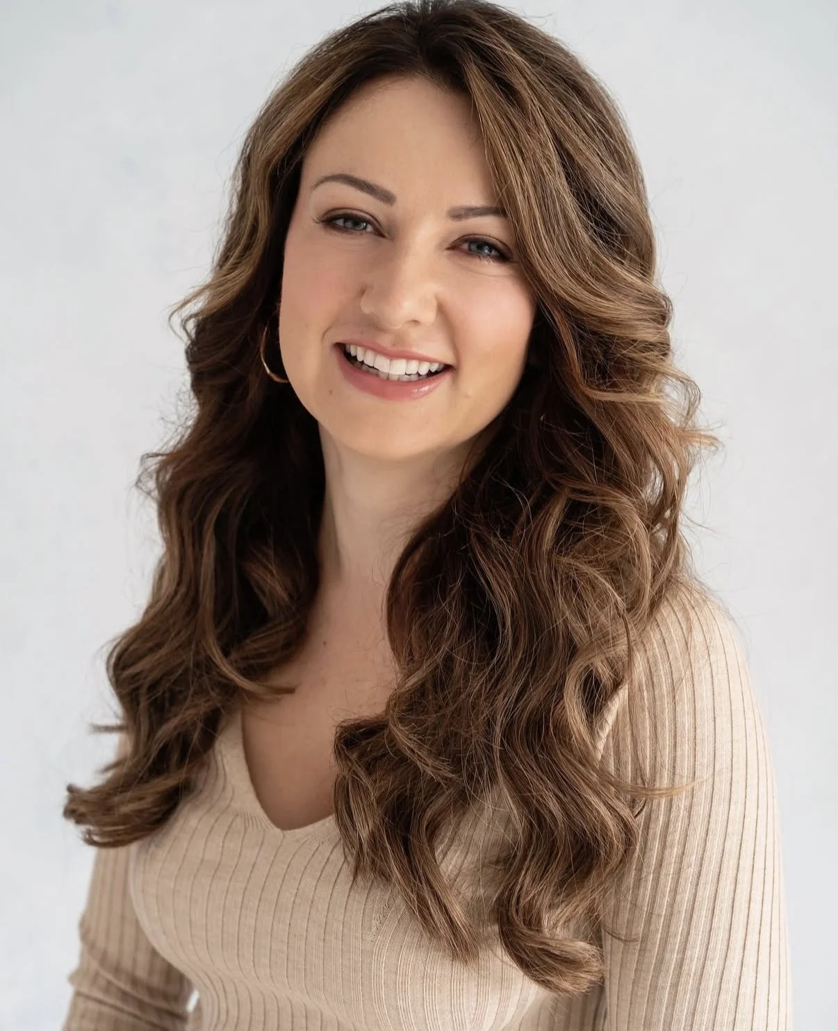 A woman with long, wavy brown hair, smiling, wearing a beige, ribbed top and hoop earrings, against a plain light background.