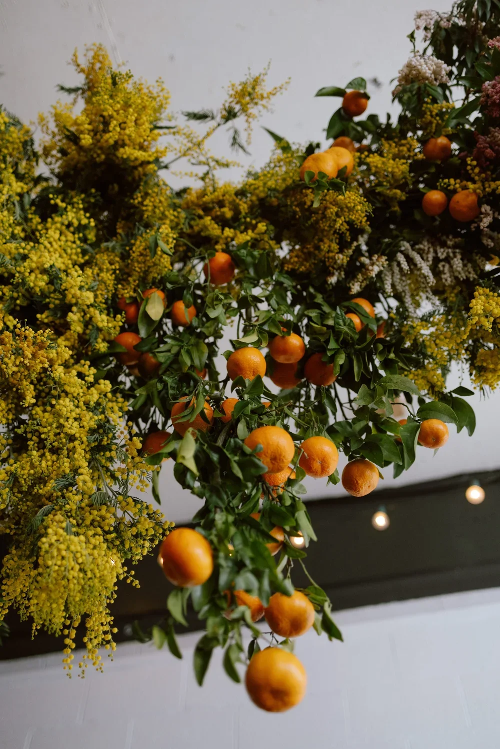 Decorative hanging arrangement of yellow flowers, green foliage, and small orange fruits, possibly tangerines or oranges, against a white background.
